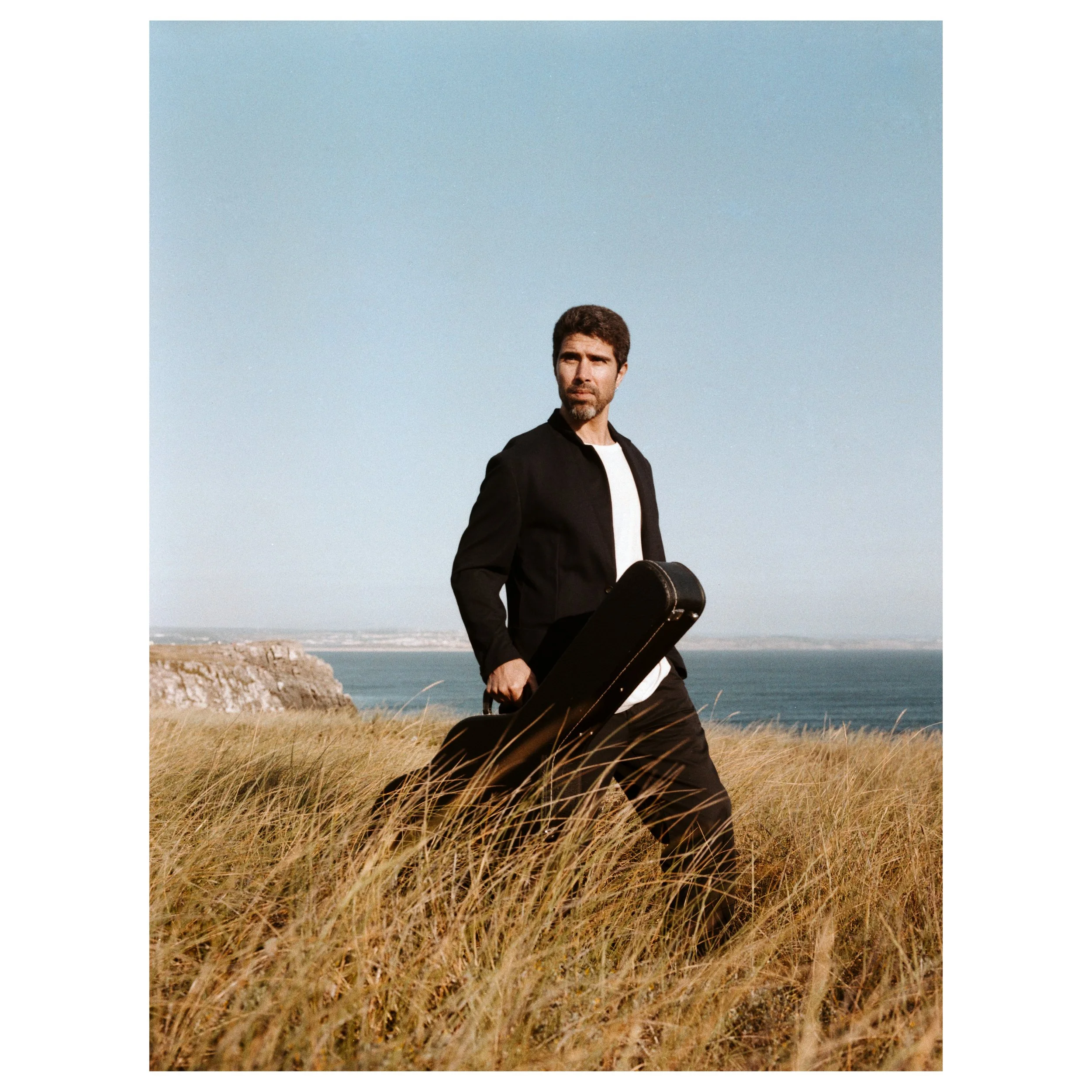 A man with dark hair and beard, dressed in a black jacket and white shirt, standing in tall grass near the coast holding a guitar case, with an ocean, cliffs, and a clear sky in the background.