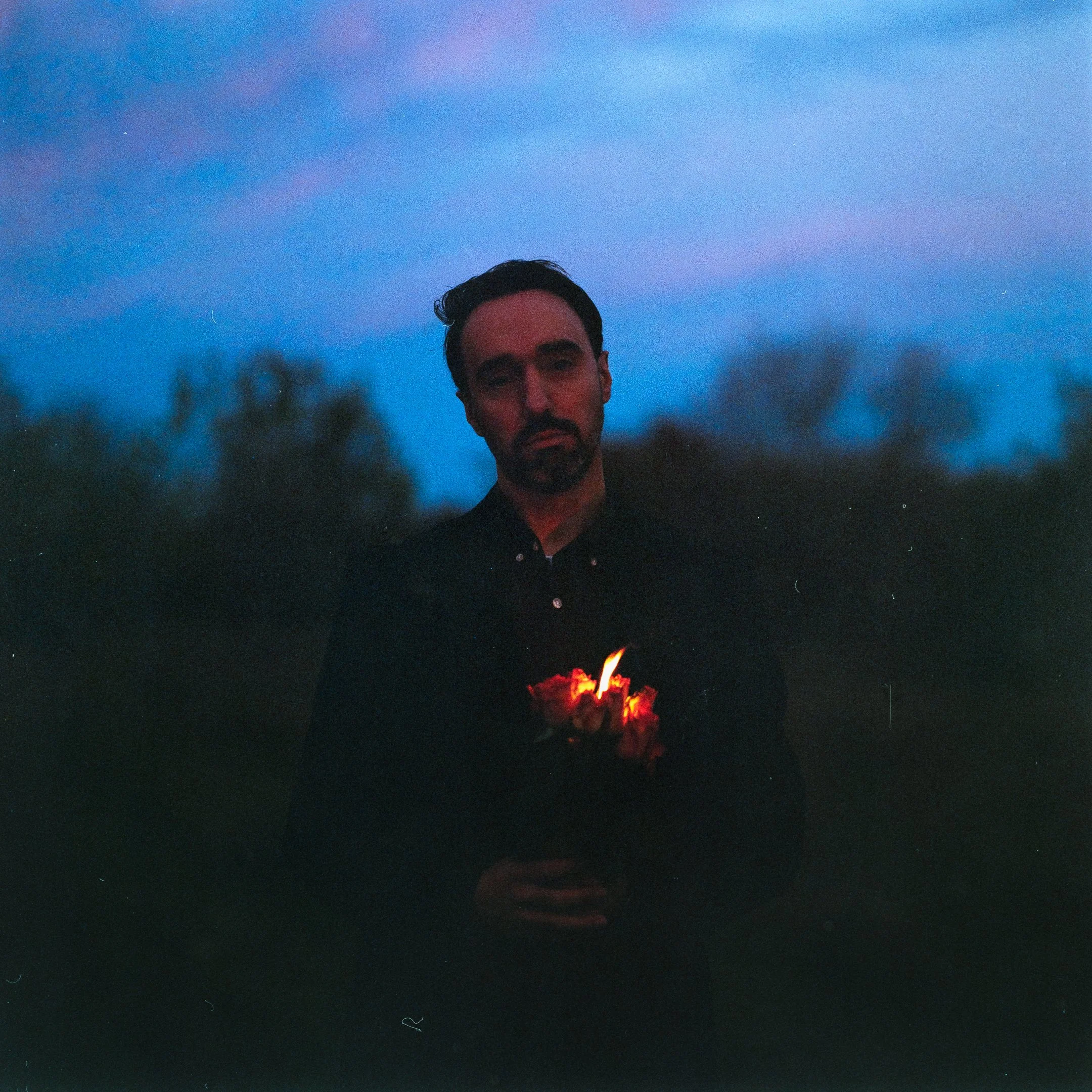 A man with dark hair and beard standing outdoors during dusk, holding a lit flare with glowing orange and red light, against a blue sky with some dark trees in the background.