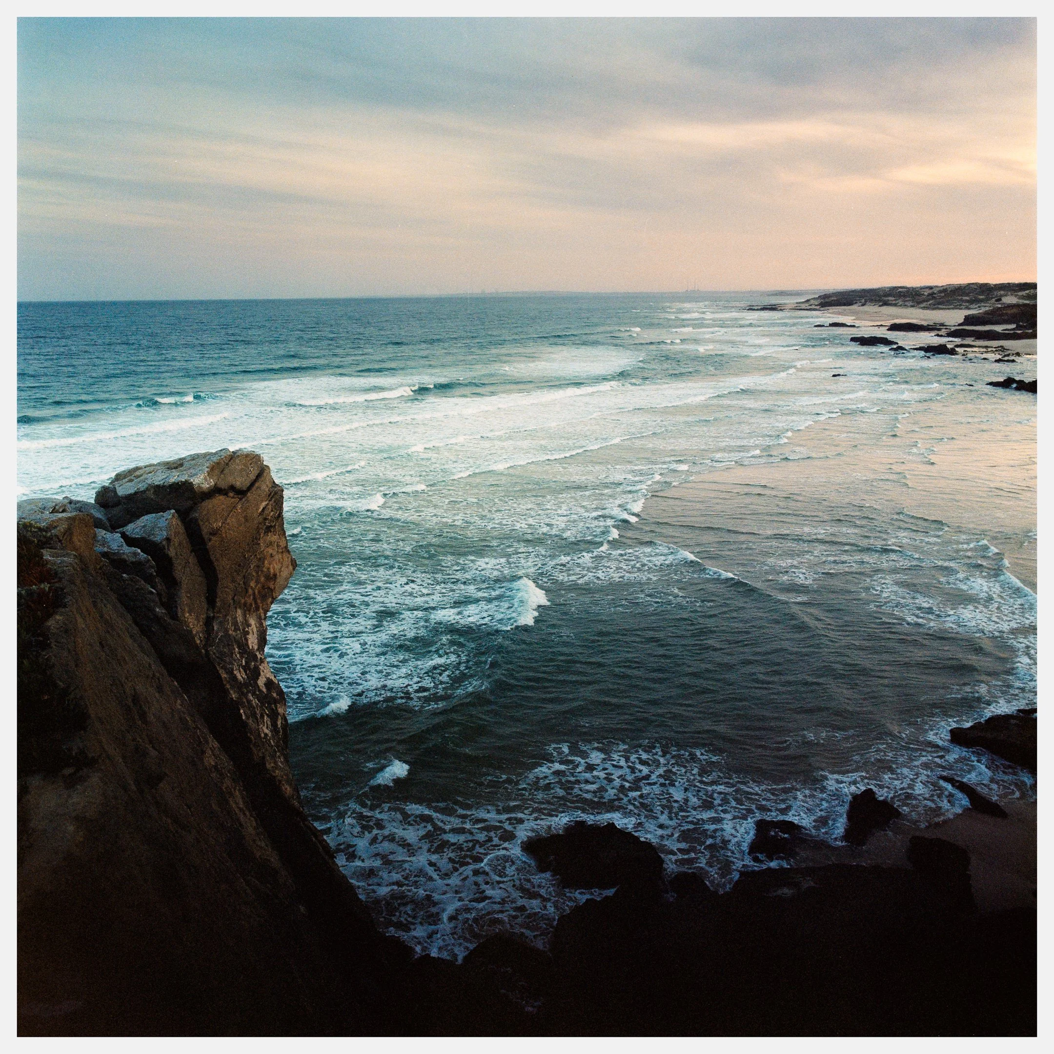 A scenic view of the ocean with waves crashing on the shore, rocky cliffs in the foreground, and a distant shoreline with gentle clouds in the sky.