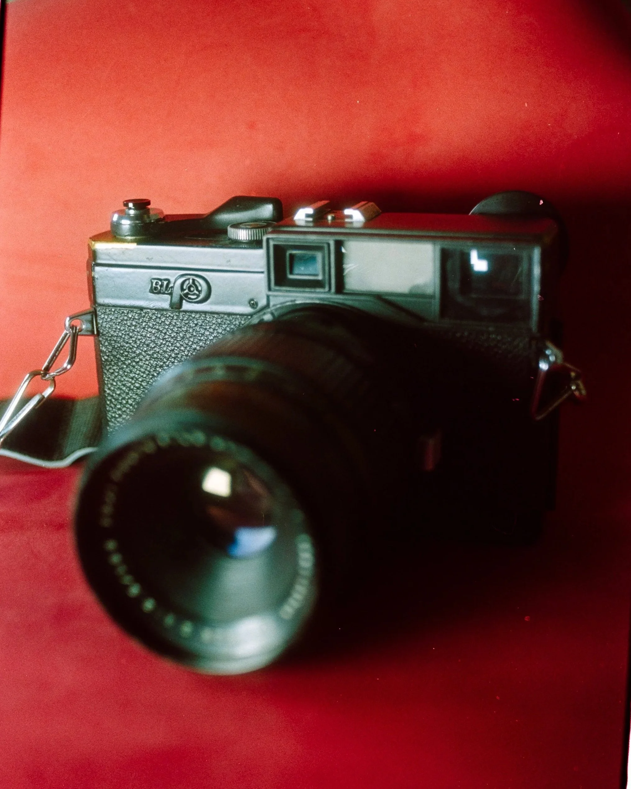 A vintage black camera with a large lens resting on a red surface, with a viewfinder and control dials visible.