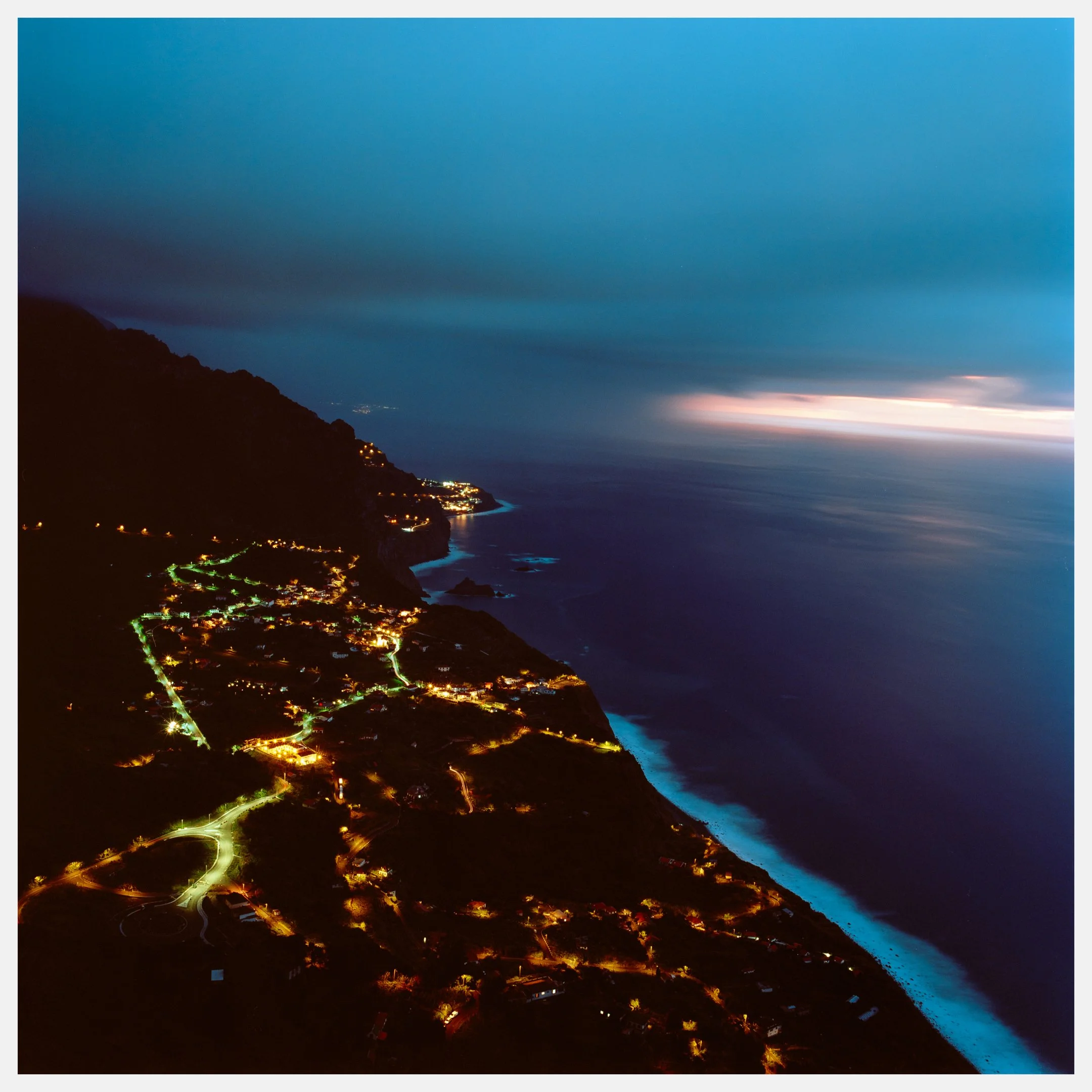 Nighttime view of a coastal town with illuminated streets along a rugged shoreline and hills, under a cloudy sky with a hint of sunset or sunrise on the horizon.
