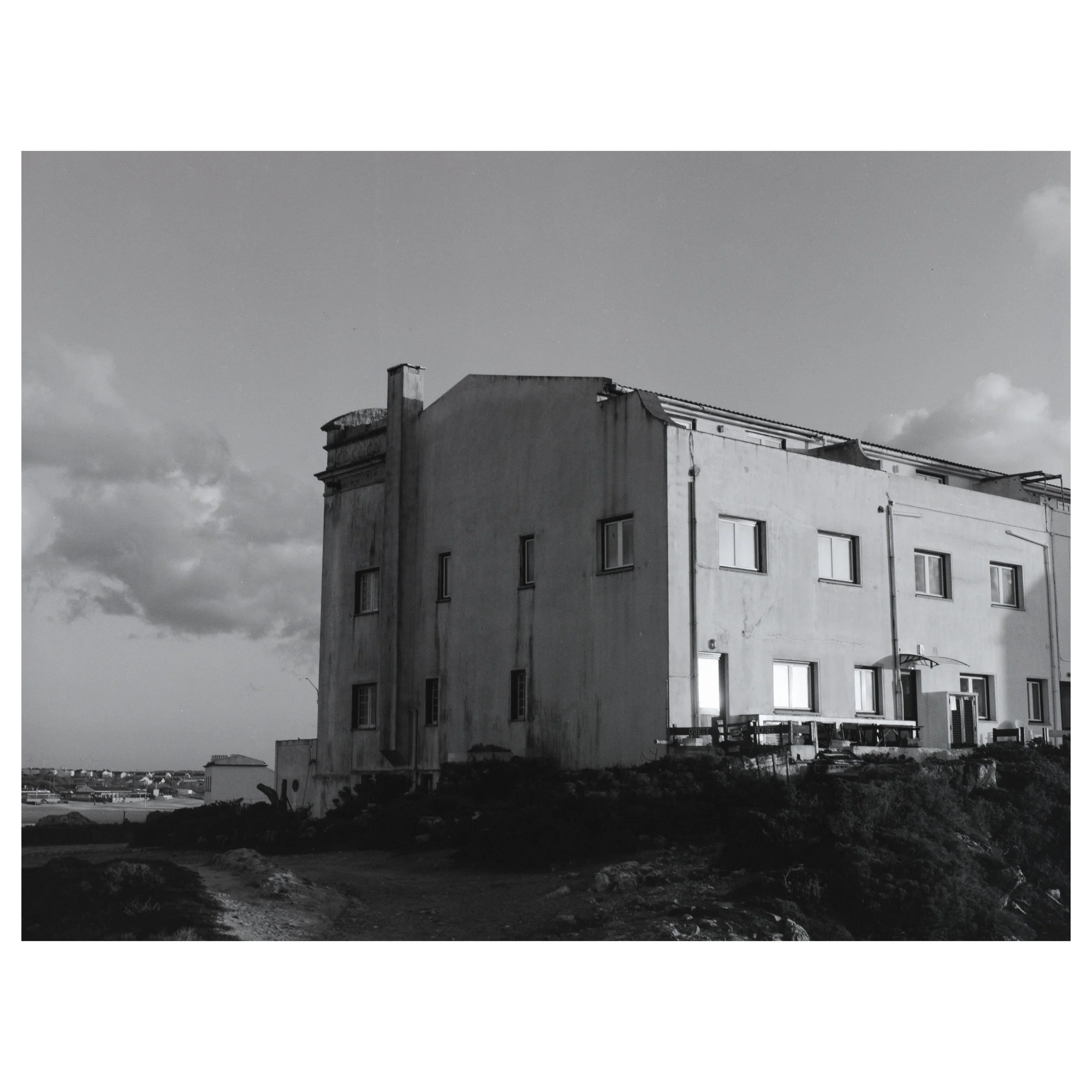 A black and white photo of a large, old, white building with multiple small windows and a flat roof, situated on uneven terrain with a dirt path in the foreground, under a partly cloudy sky.