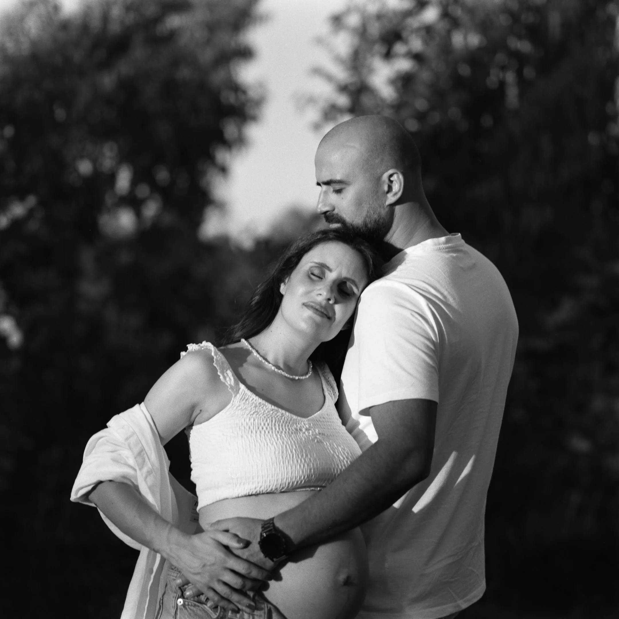 A black-and-white photo of a pregnant woman and a man embracing outdoors, with the woman resting her head on the man's shoulder and holding her belly, while the man holds her waist and kisses her forehead.