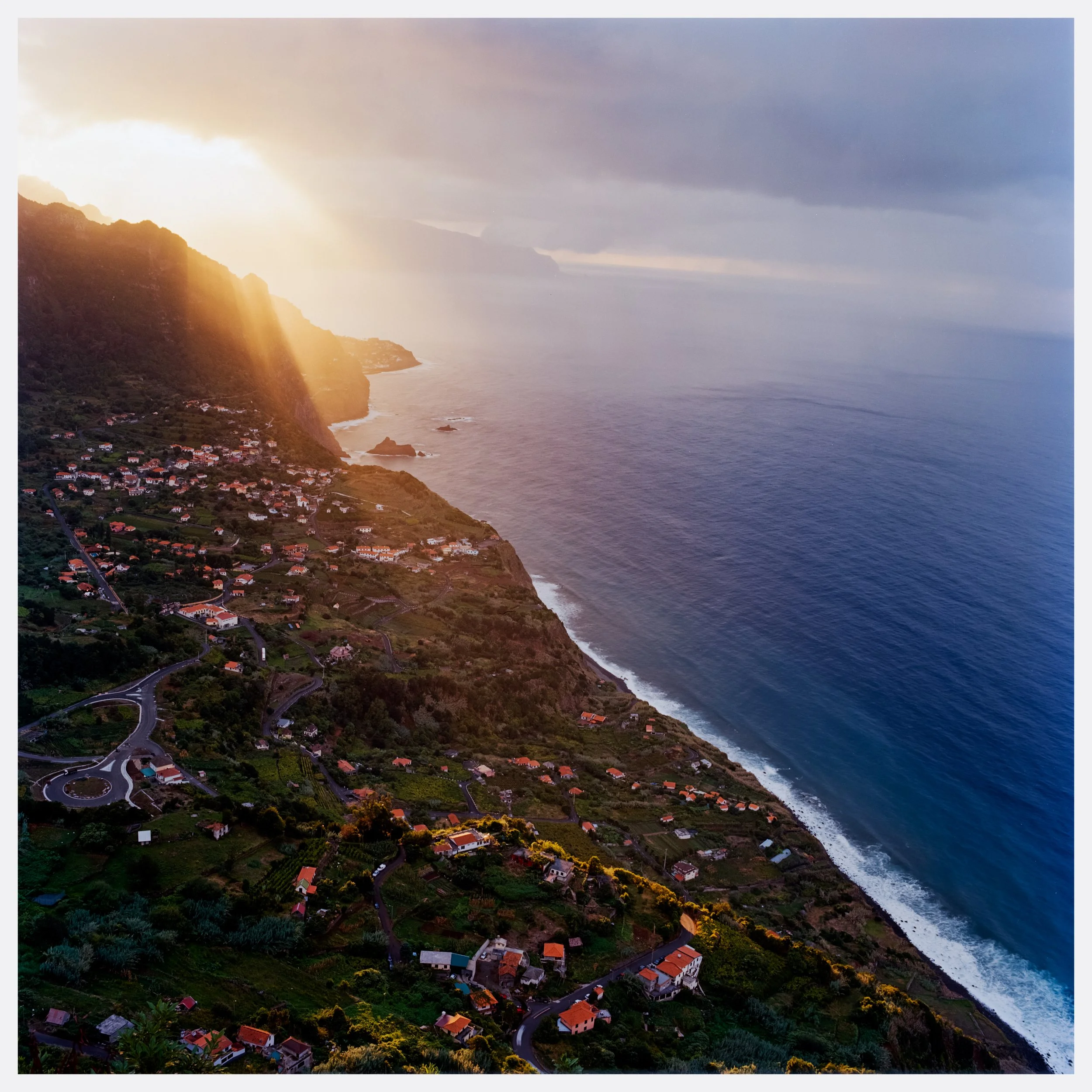 A coastal landscape with cliffs, ocean, and a small town with houses and winding roads at sunset.