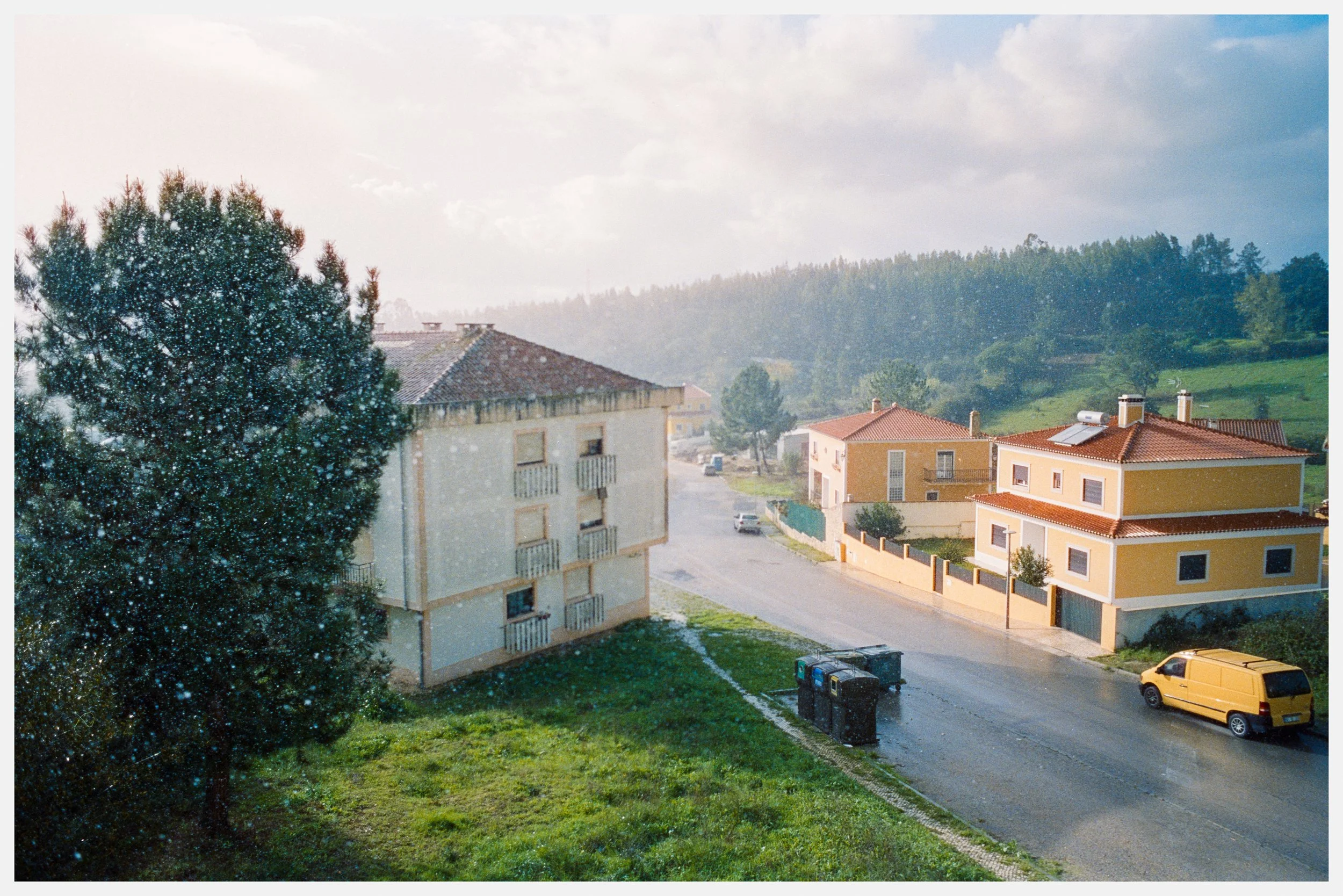 Rain falling on a suburban neighborhood with a tree in the foreground, a beige apartment building, and yellow houses with red roofs, parked cars, and trash bins, under a cloudy sky during daytime.