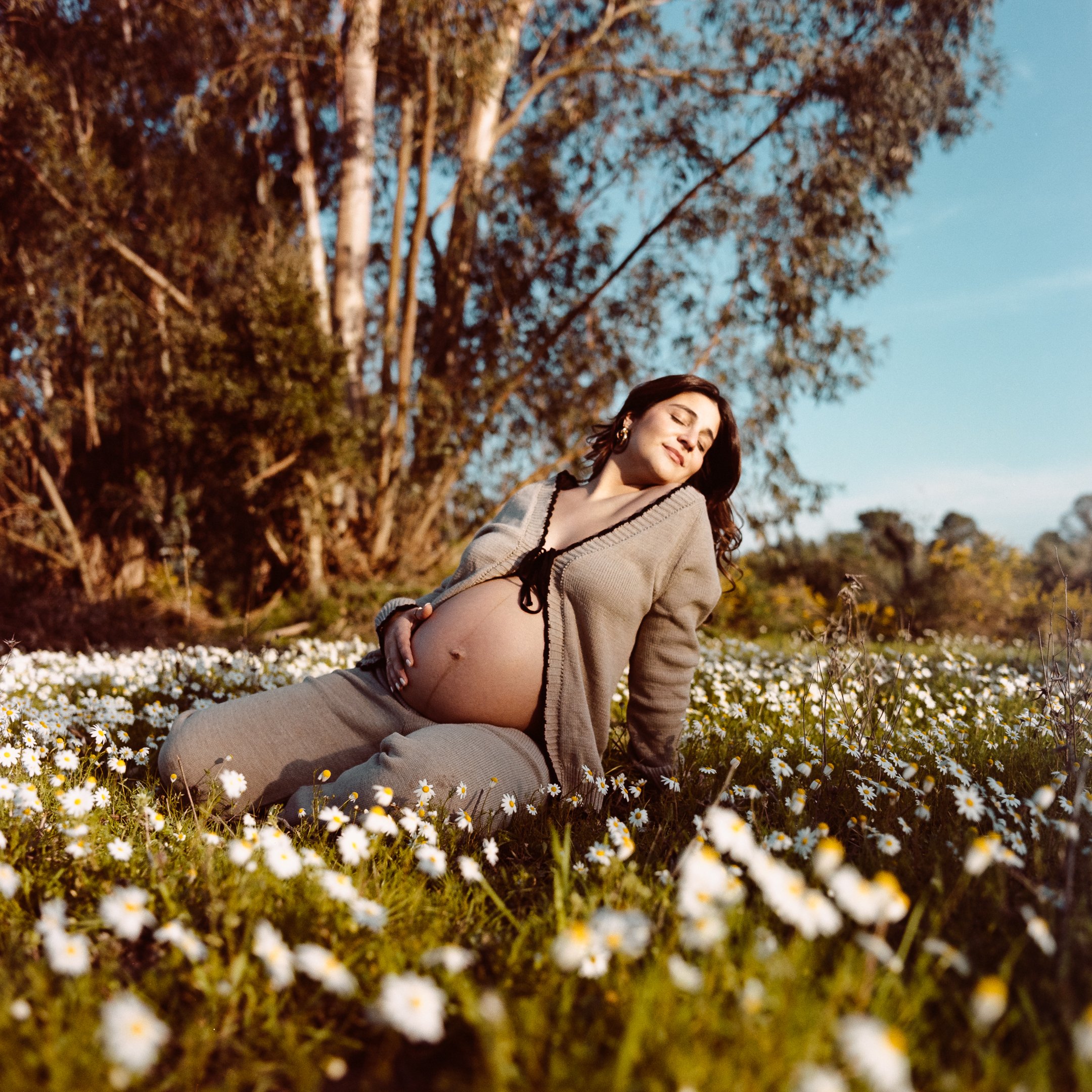 Pregnant woman sitting in a field of daisies, smiling with eyes closed, wearing a beige cardigan and matching pants, with a backdrop of trees and a blue sky.