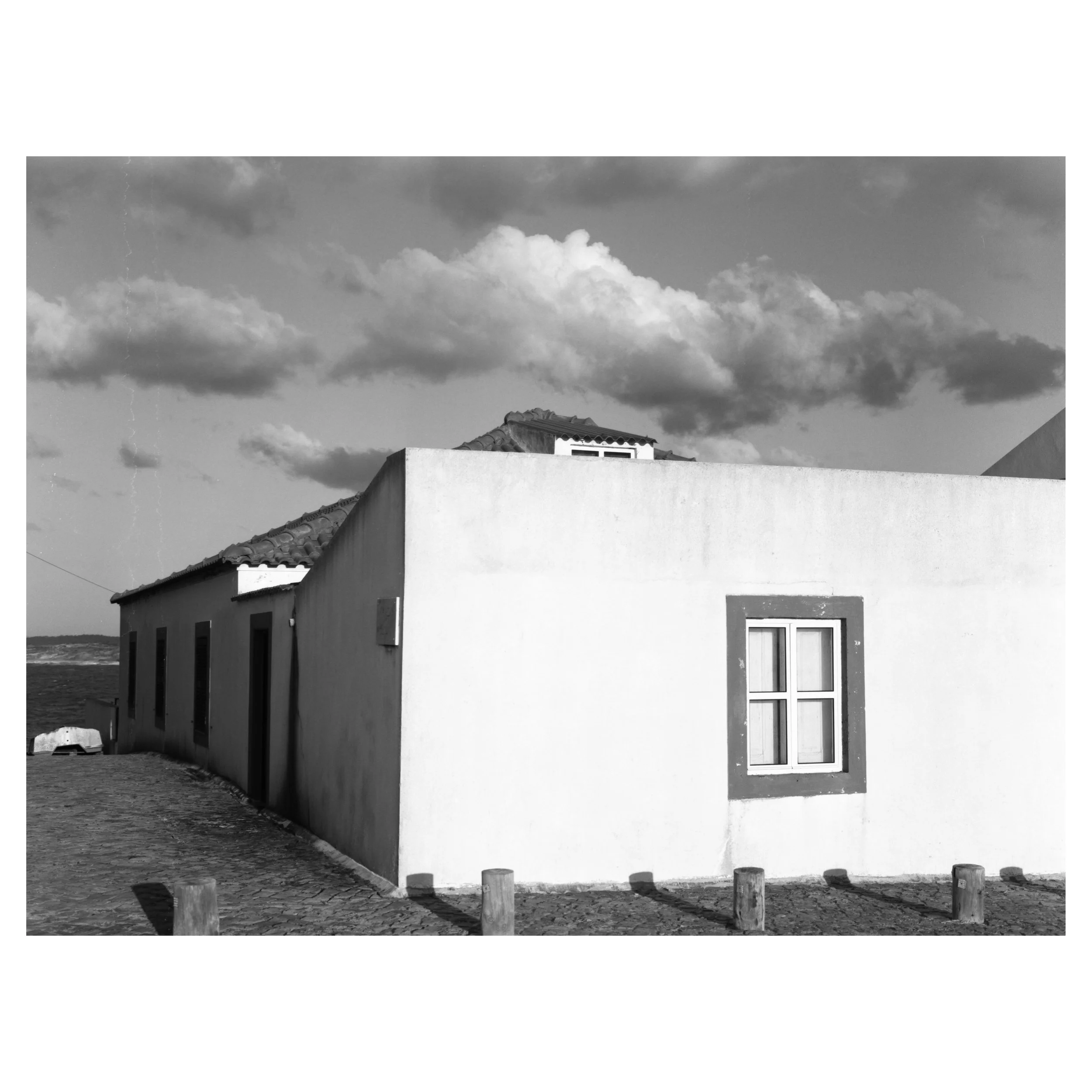 A black and white photo of a building with a flat wall and a window, with a cloudy sky in the background.