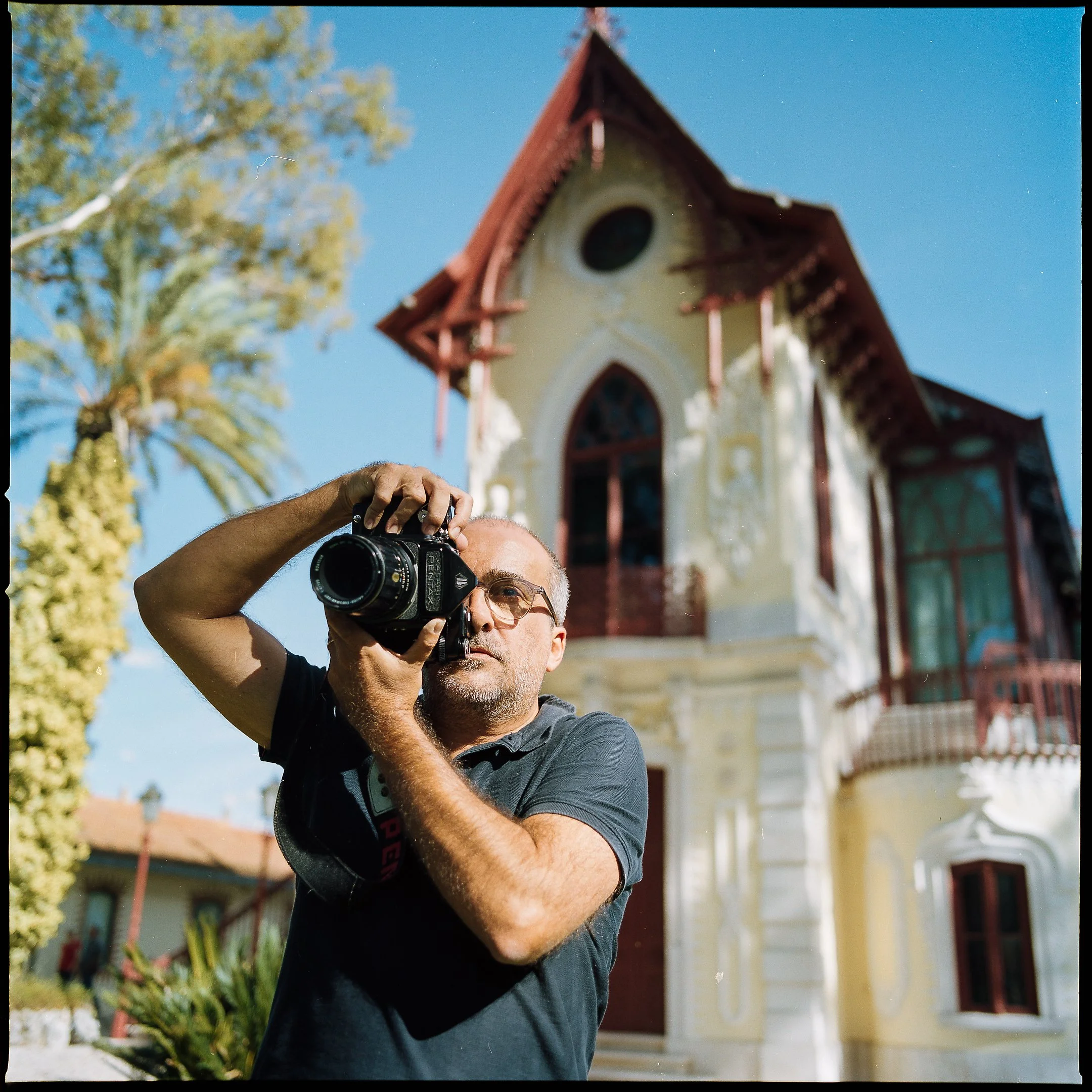 A man with gray hair, glasses, and a dark shirt taking a photograph with a camera, standing in front of a colorful, historic house with a steep, pointed roof, surrounded by trees and plants on a sunny day.