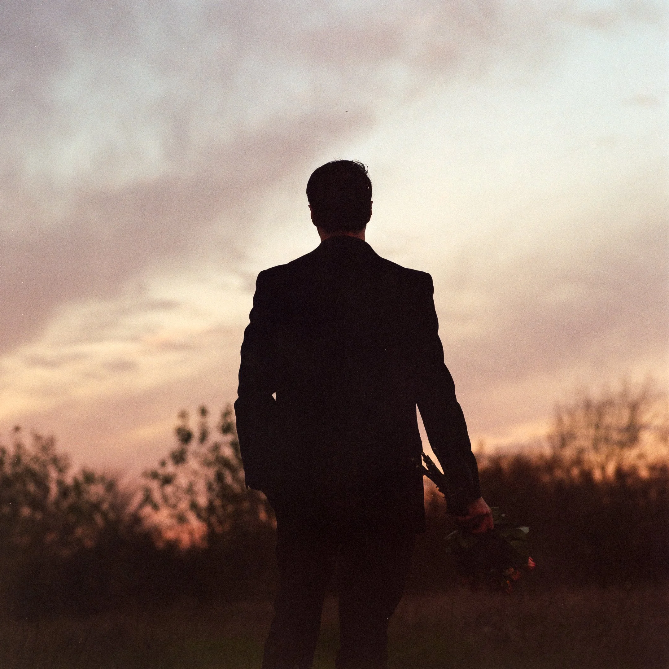 Silhouette of a man in a suit holding a bouquet of flowers, standing outdoors at sunset with a cloudy sky and trees in the background.