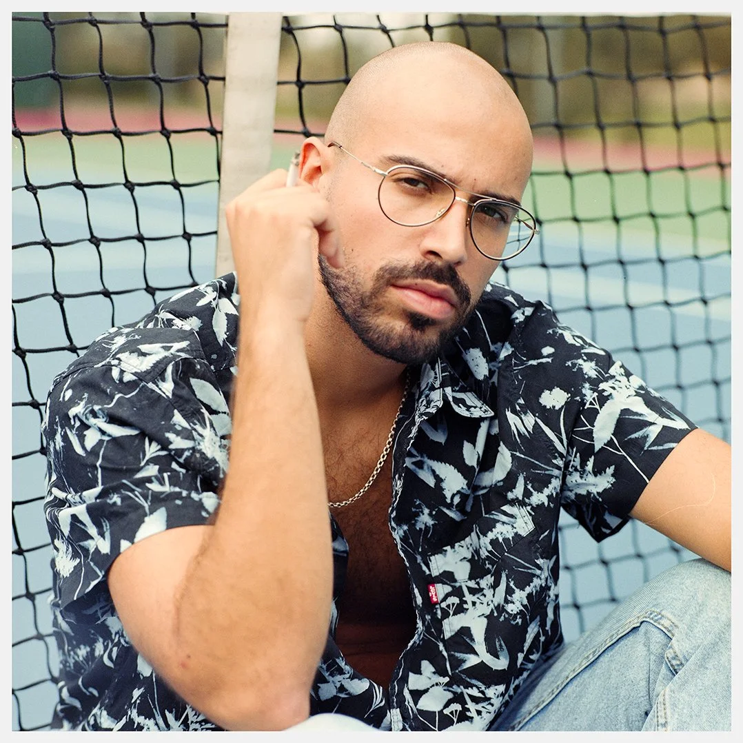 A man with a shaved head, glasses, and a goatee sitting near a tennis court fence, wearing a black and white patterned shirt and a chain necklace, looking at the camera.