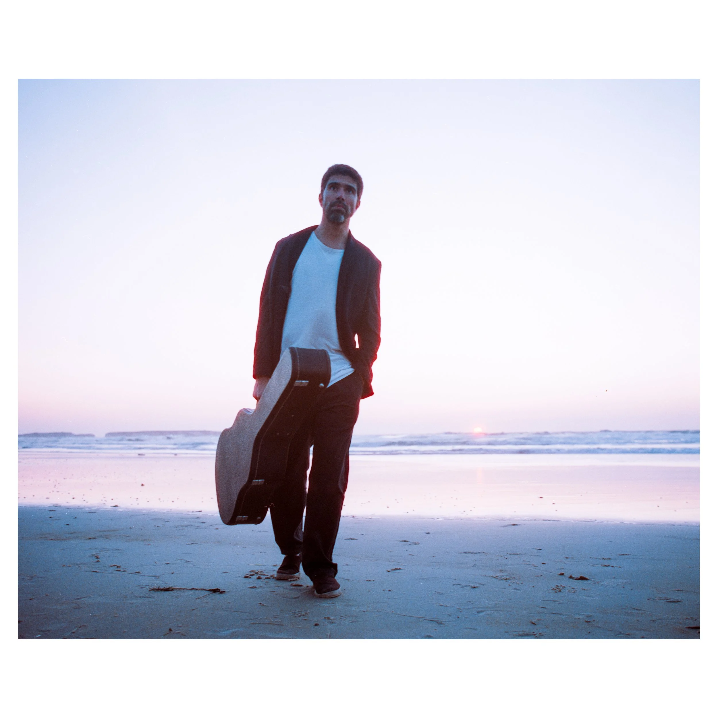 A man walking on the beach at sunset, carrying a guitar case. photo by Bruno Novais
