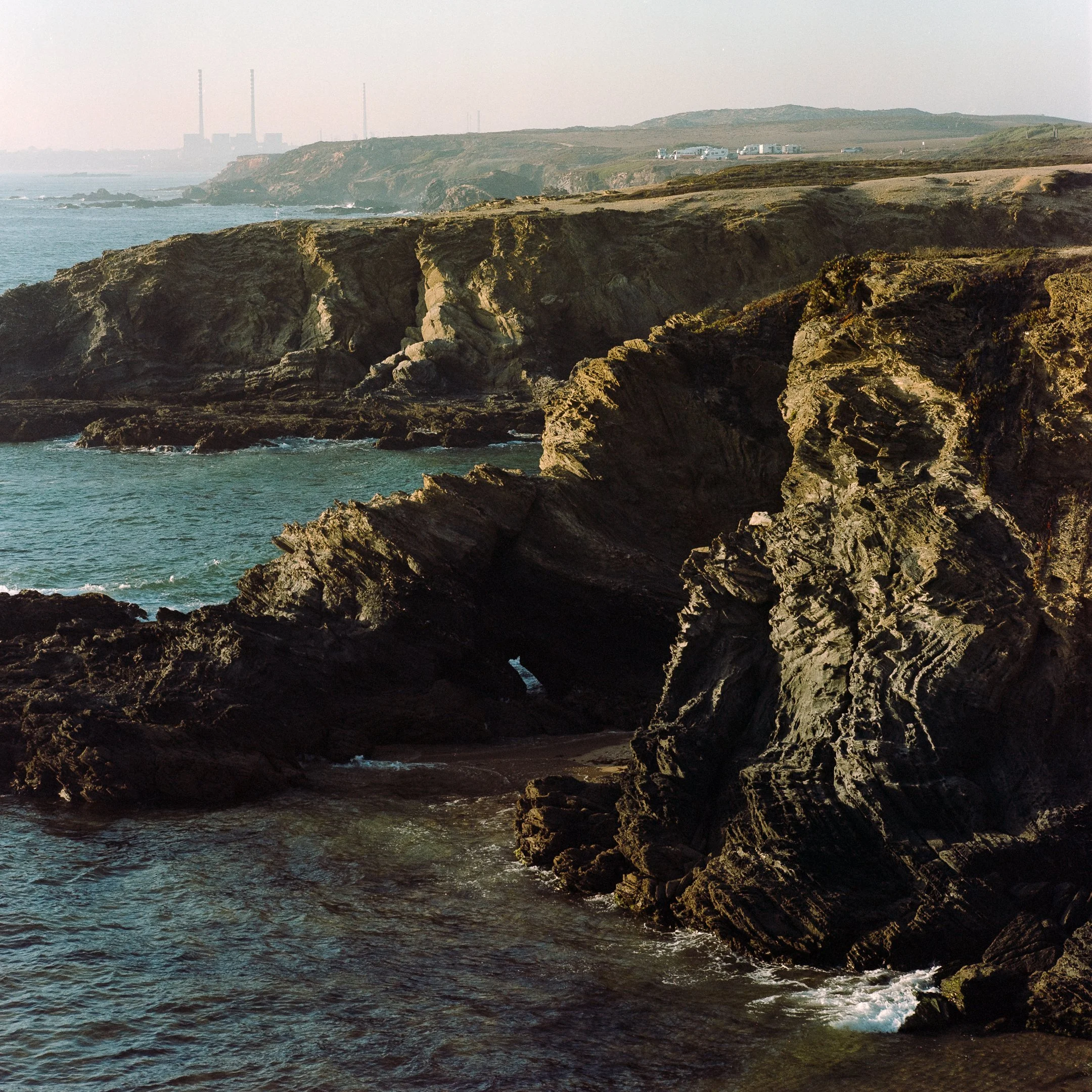 Rocky coastal cliffs and ocean waves with industrial buildings and smokestacks in the background.
