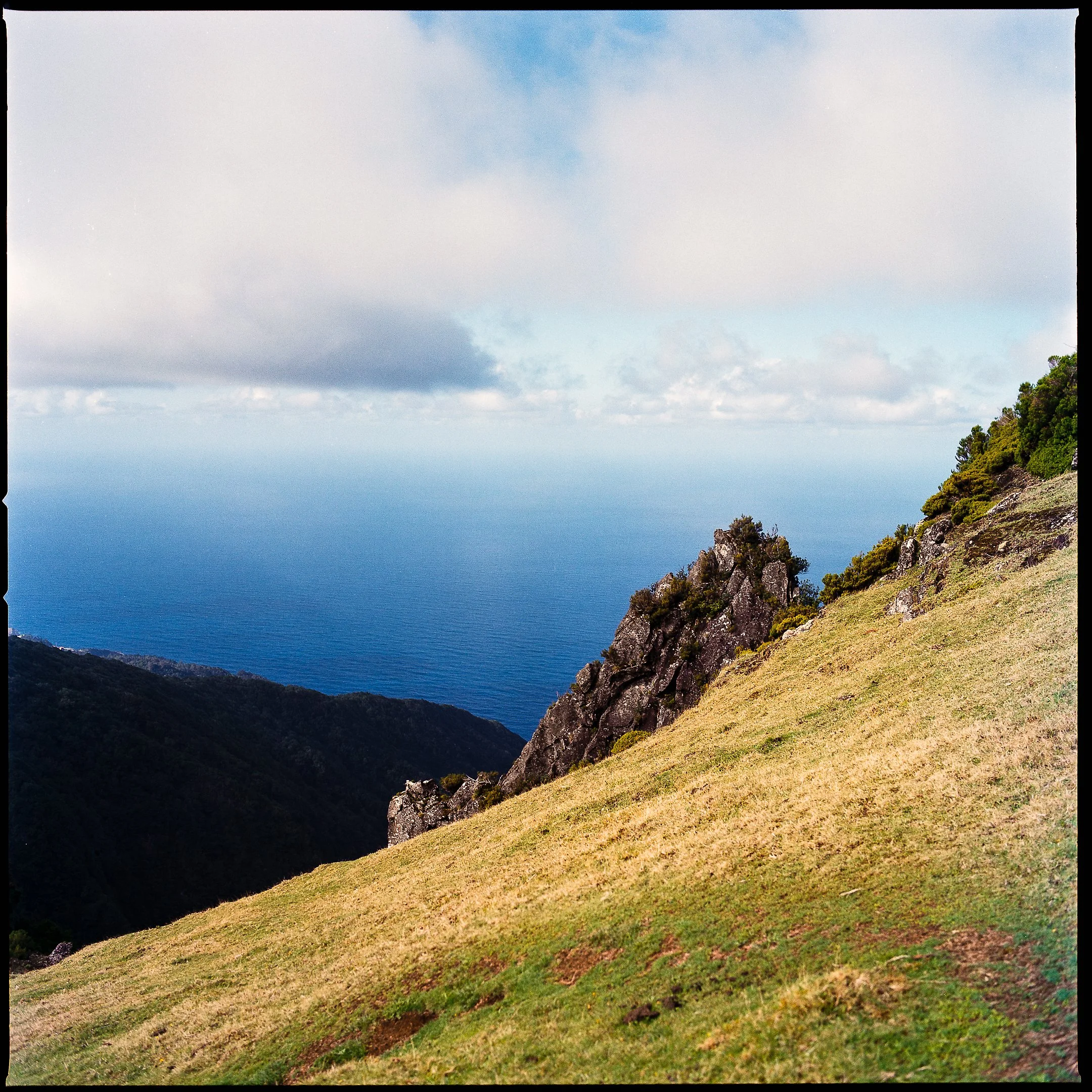 A hillside with green grass and rocks, overlooking a deep blue ocean under a cloudy sky.