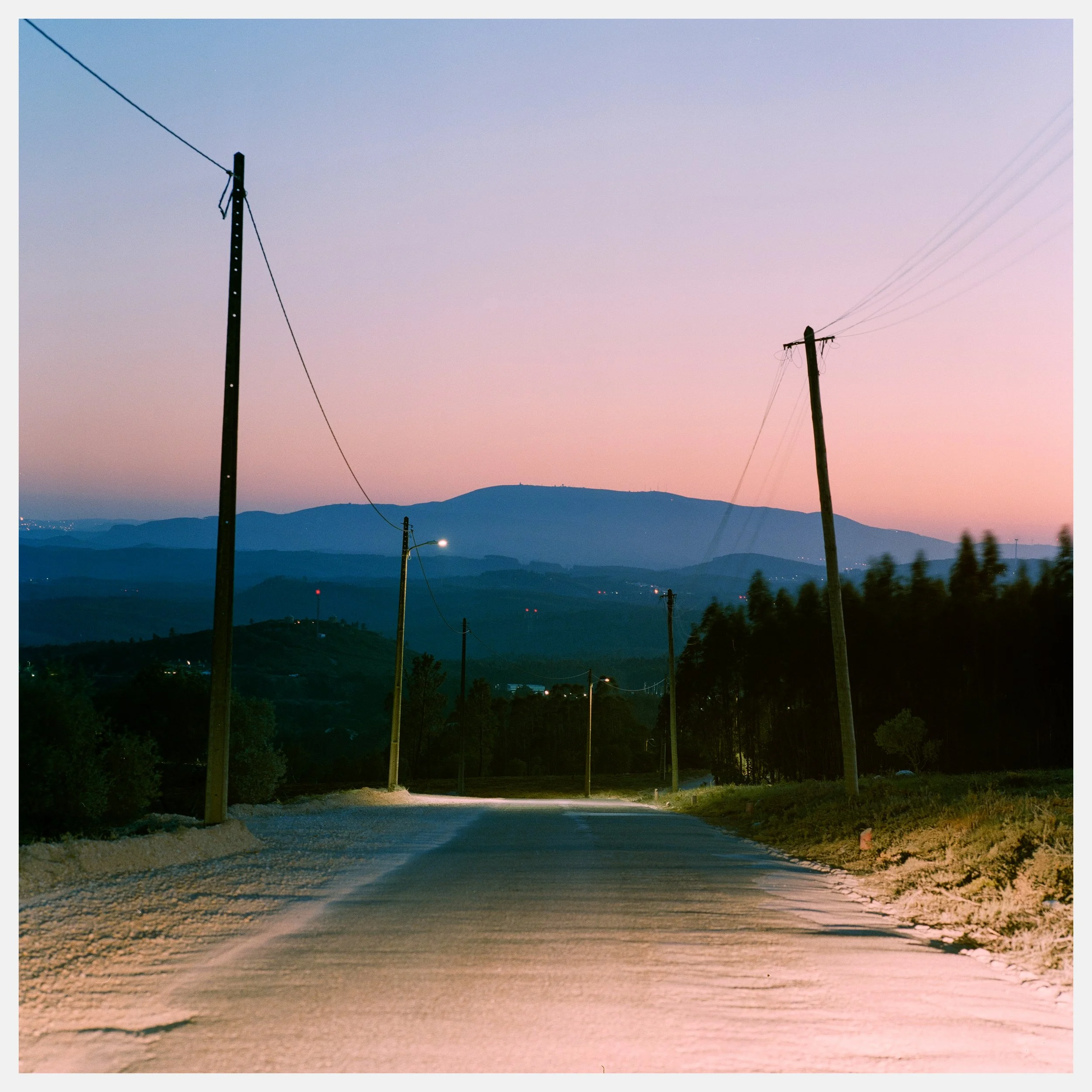 A rural road at dusk with utility poles and power lines stretching into the distance, hills and mountains in the background, and a sky transitioning from pink to blue.