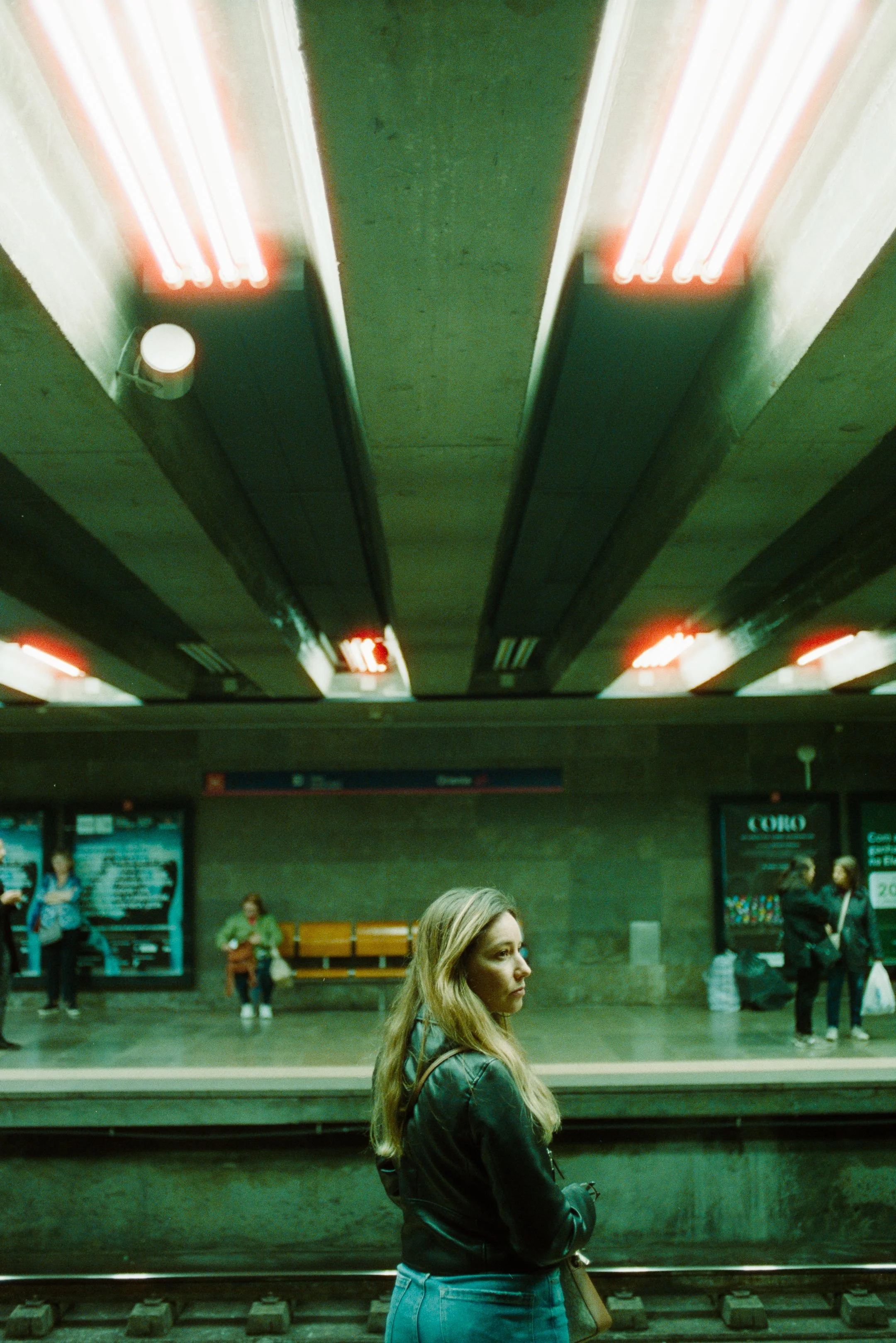 A woman with long blonde hair wearing a black jacket and jeans waiting at a subway station, with other travelers in the background and illuminated ceiling lights above. photo by Bruno Novais