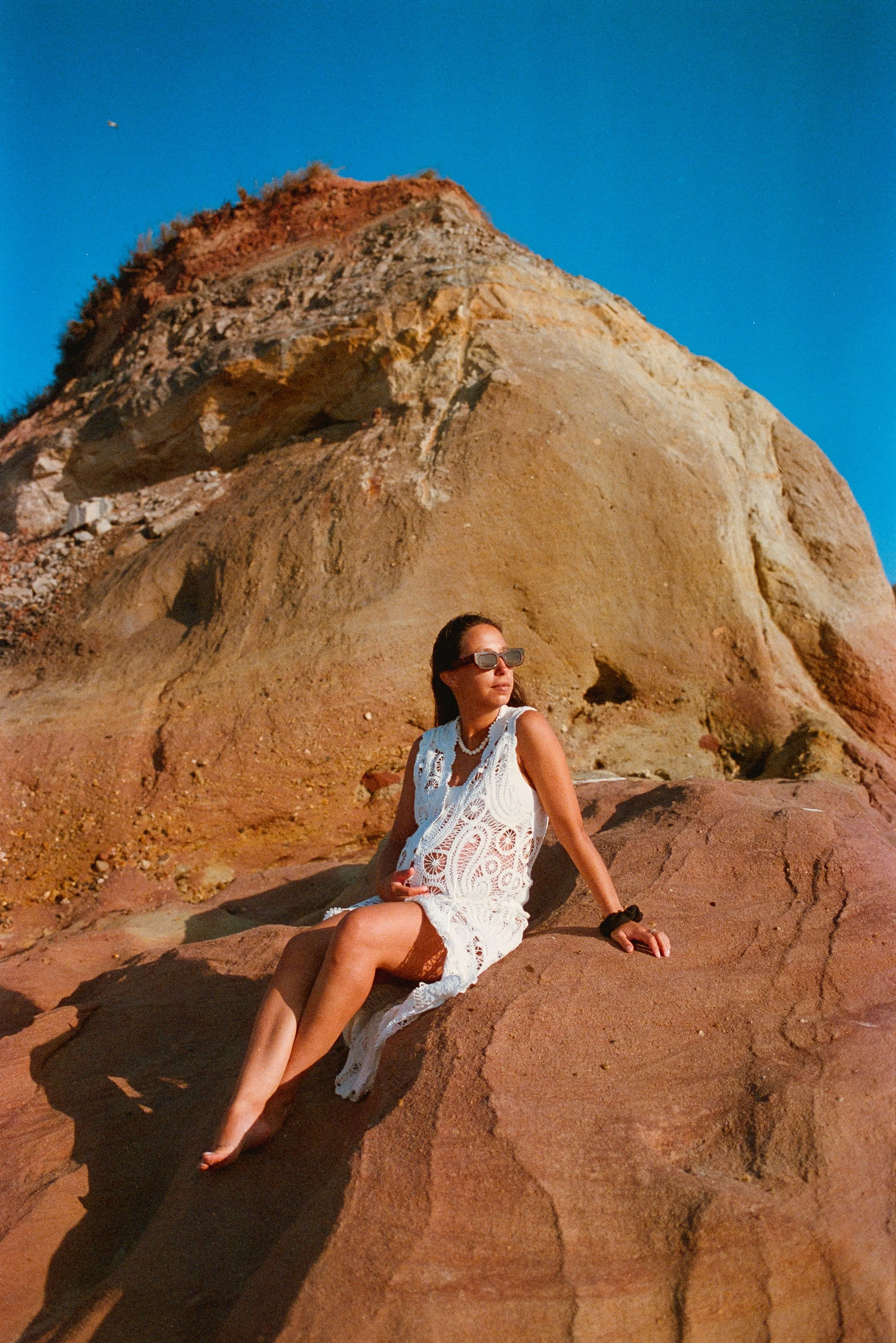 A woman in sunglasses sitting on reddish rocks with a large, colorful hill or mountain behind her against a clear blue sky.