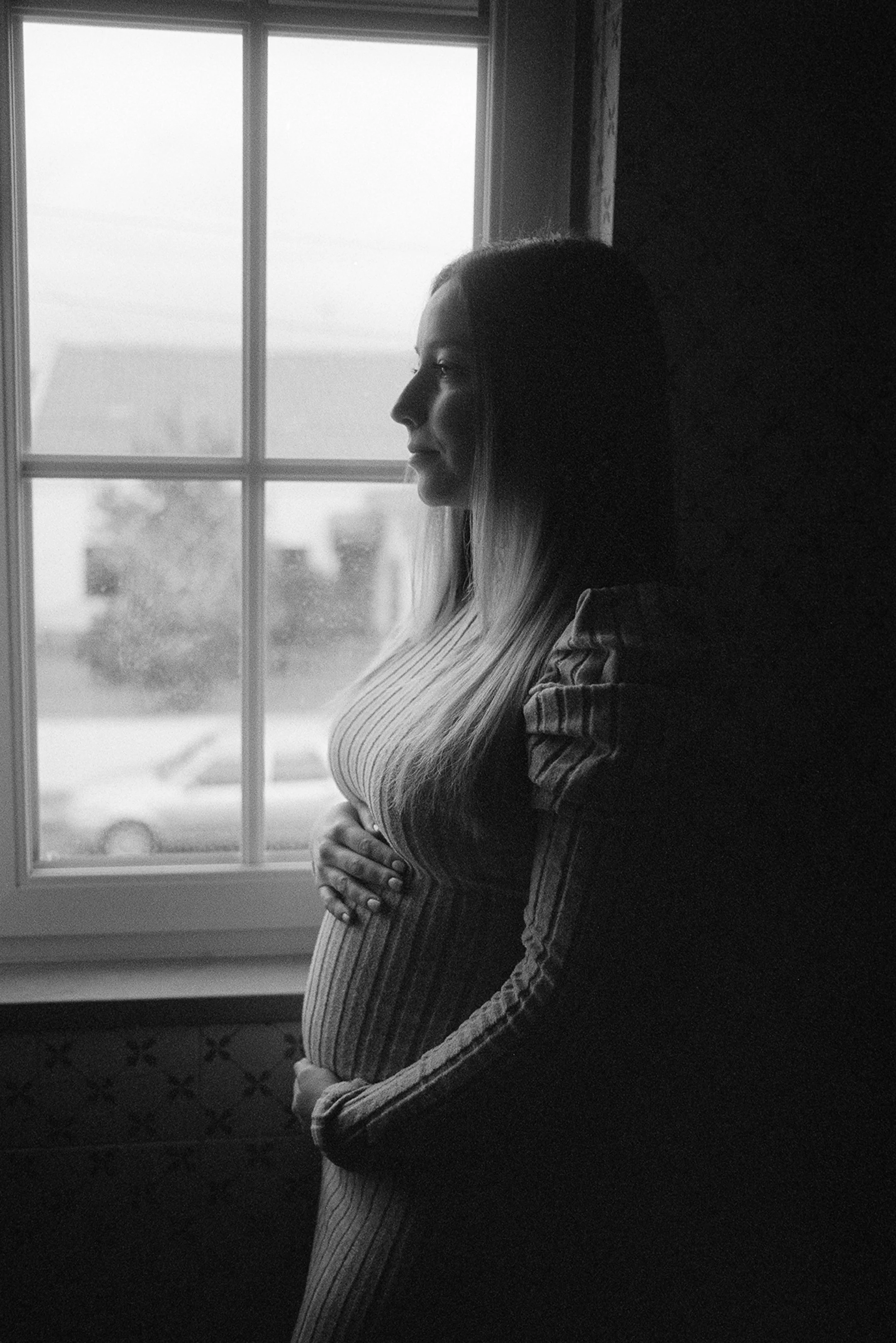 Black and white photo of a pregnant woman standing by a window, looking outside with her hands resting on her belly.