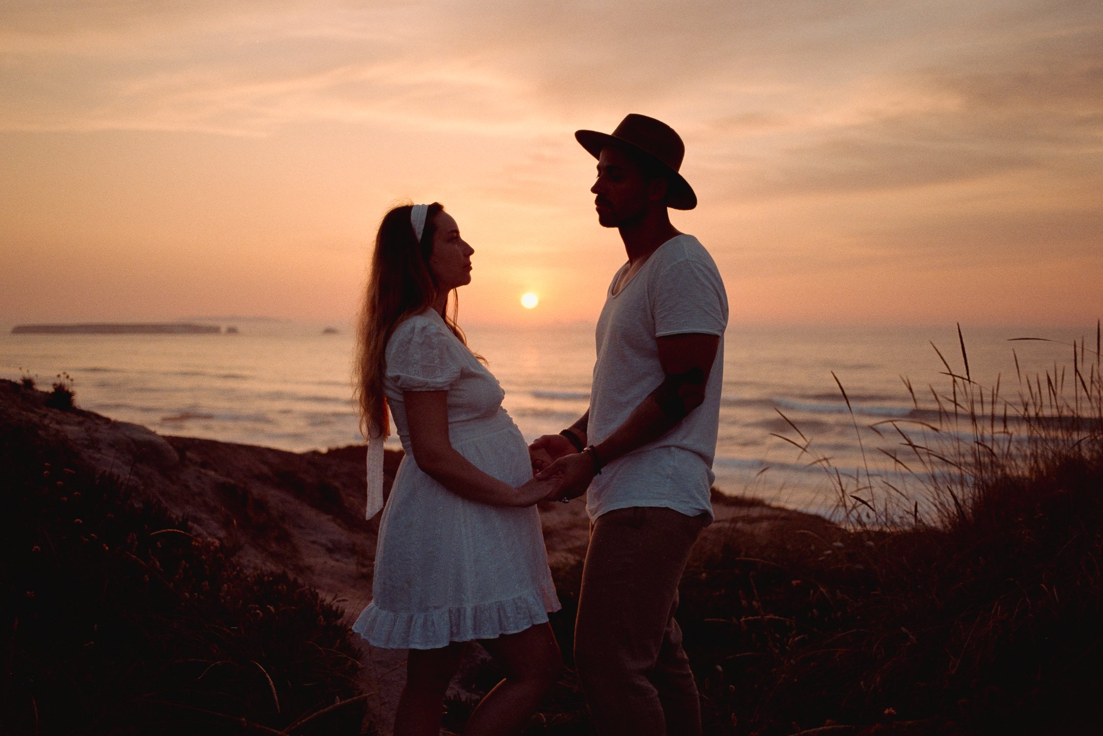 A pregnant woman and a man holding hands and looking at each other during sunset on a beach.