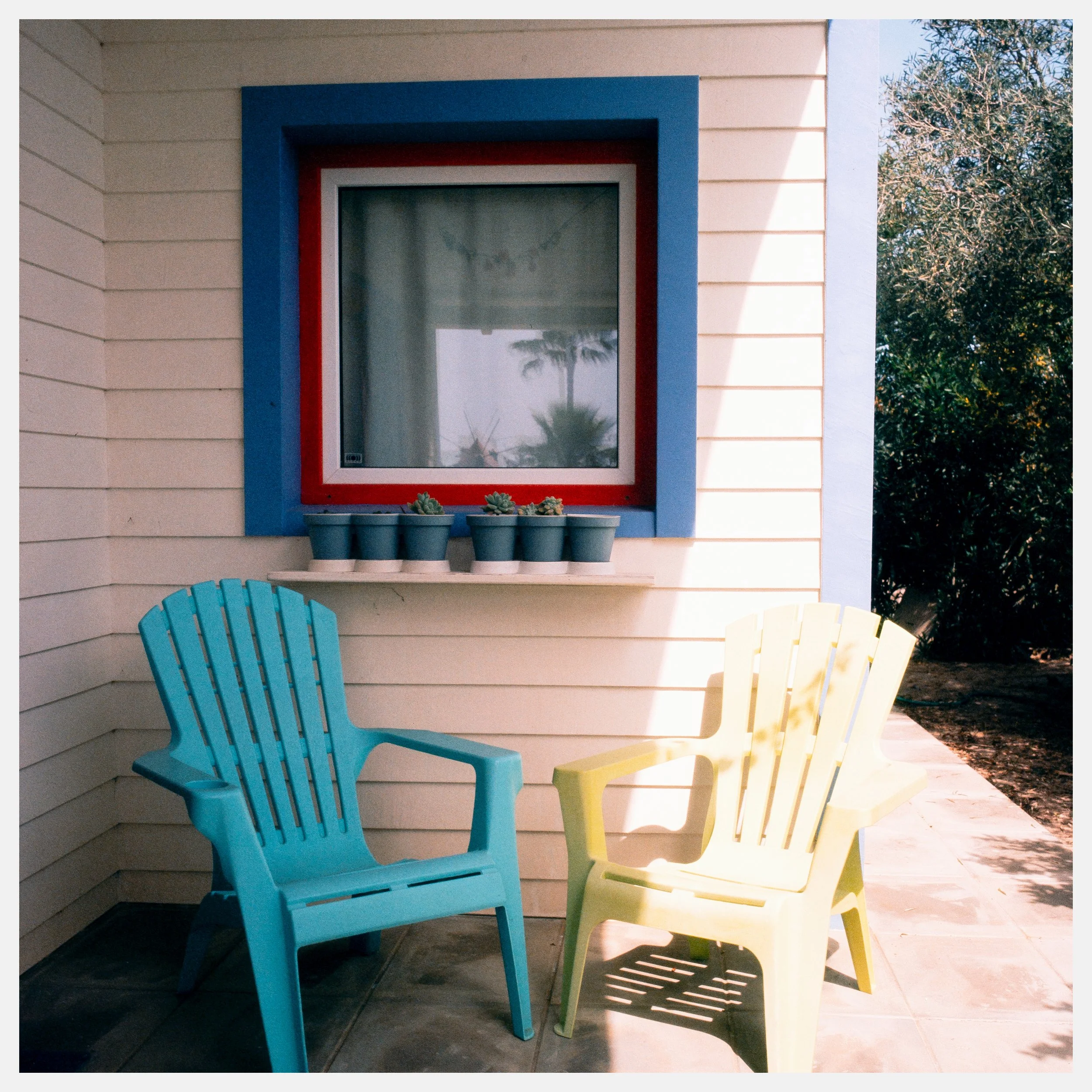 Colorful backyard patio with blue and yellow Adirondack chairs, window with red and blue frame, and potted cacti on bench