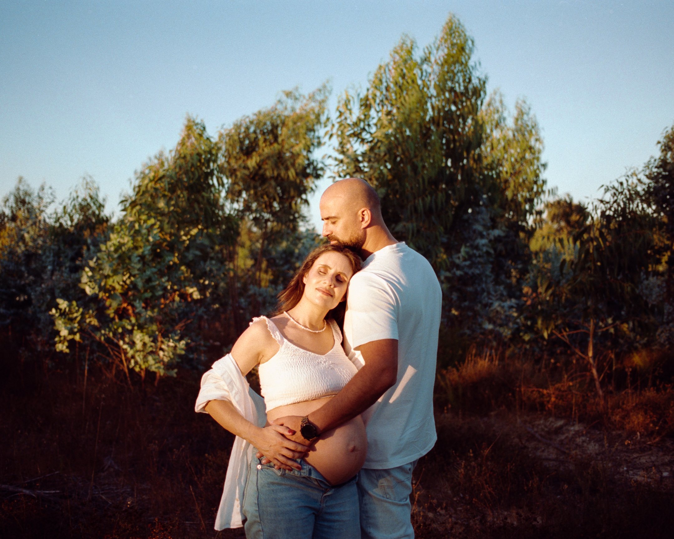 A pregnant woman and her partner standing outdoors during sunset, embracing lovingly with trees in the background.