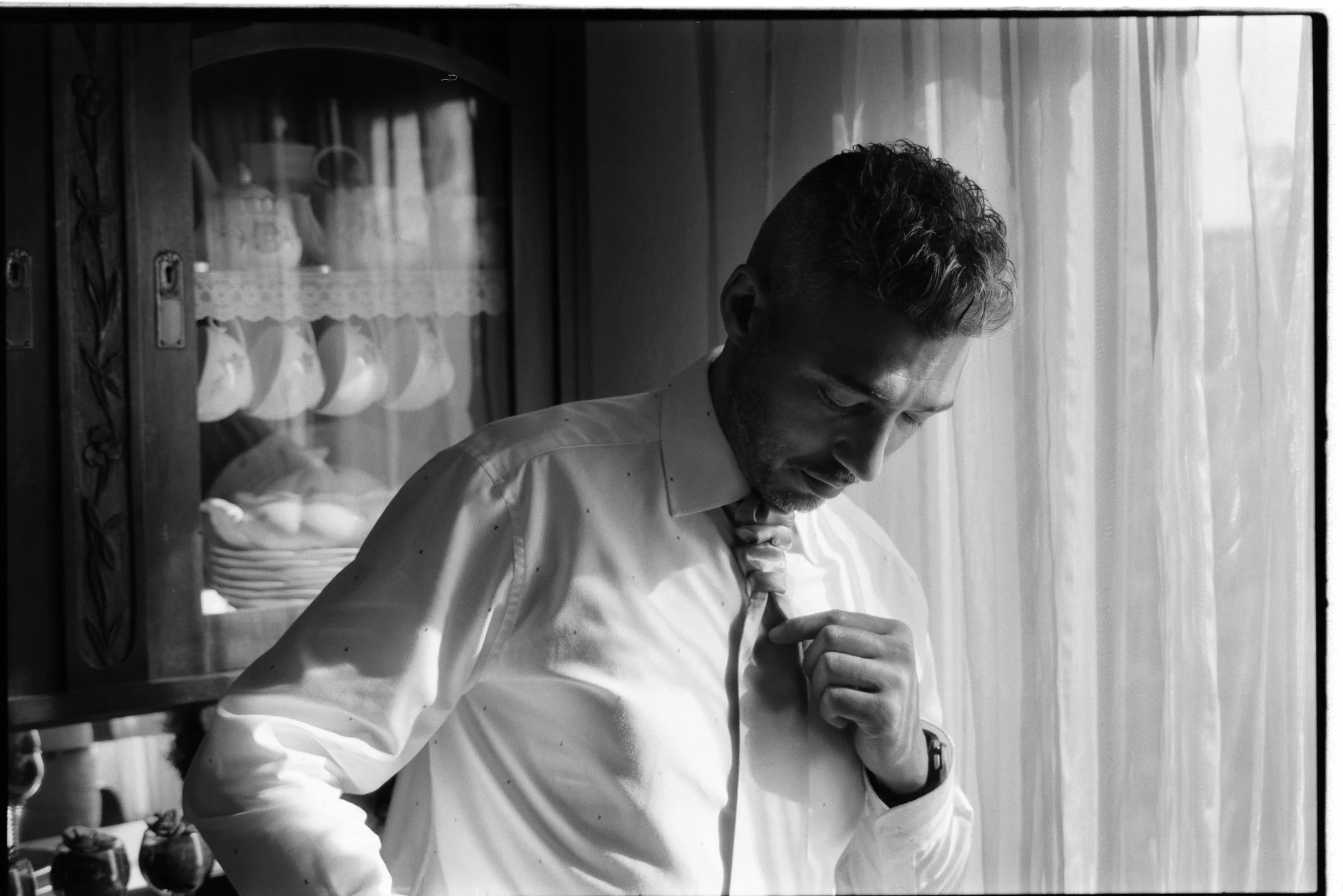 A man adjusting his tie while standing by a window with curtains, with a cabinet displaying dishes in the background. photo by Bruno Novais