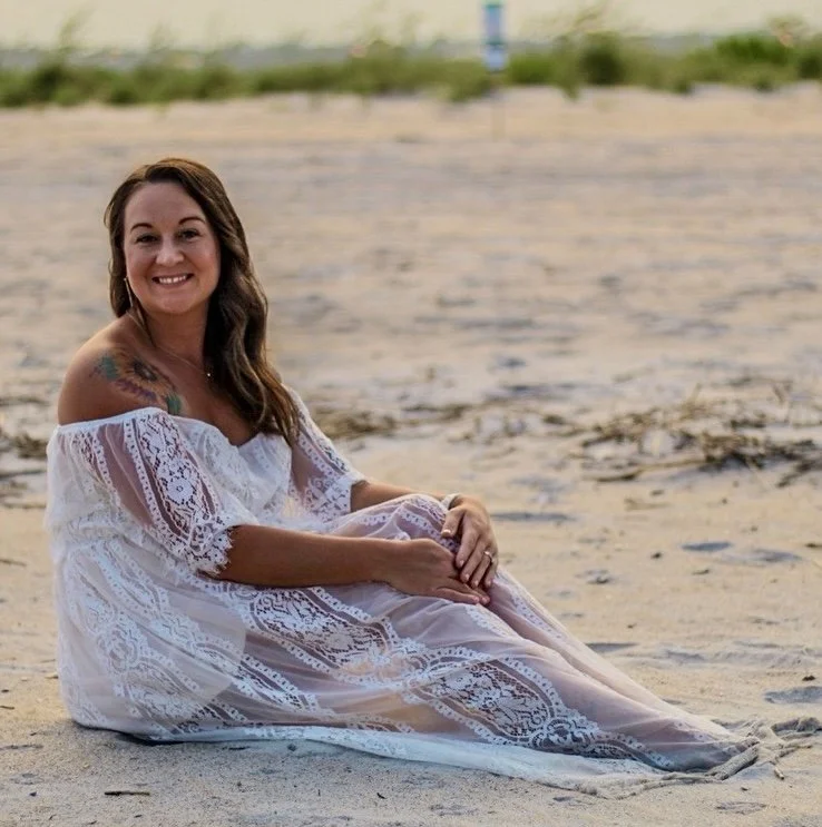 Woman sitting on a sandy beach wearing a white lace off-the-shoulder dress, smiling at the camera.