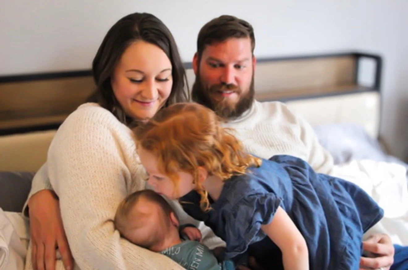 A family of five sitting on a bed, with two adults and three children, including a baby, enjoying a moment together.