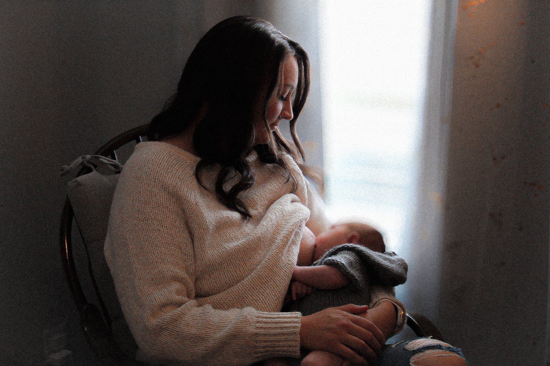 A woman with long, dark, wavy hair sitting in a softly lit room, breastfeeding a baby while smiling gently.