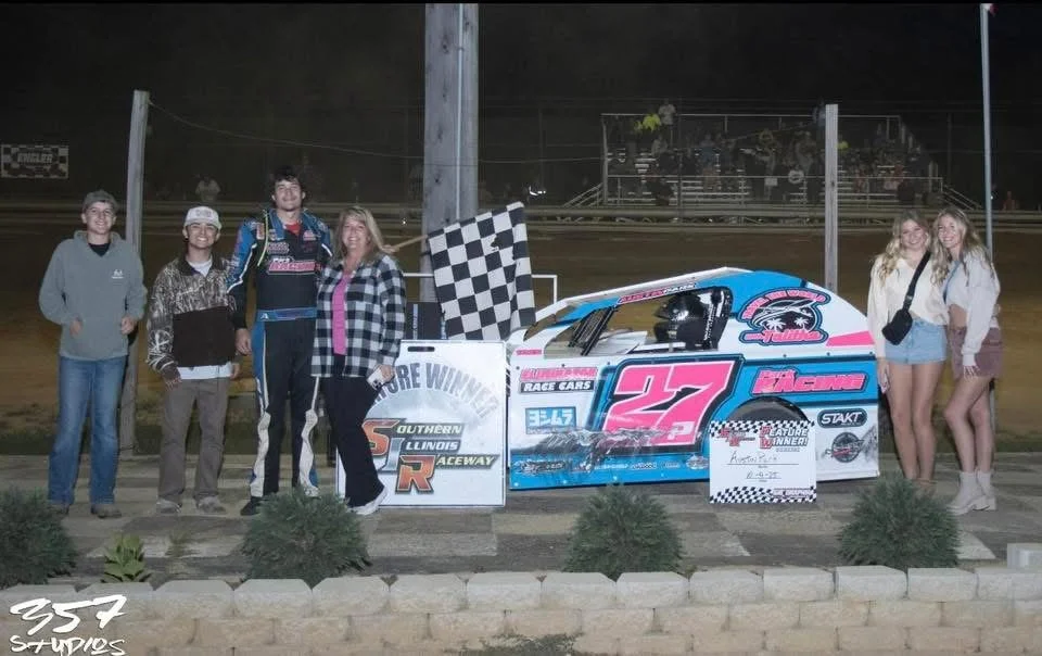Group of people celebrating a race car victory at a dirt raceway, with checkered flags and winners' signs.