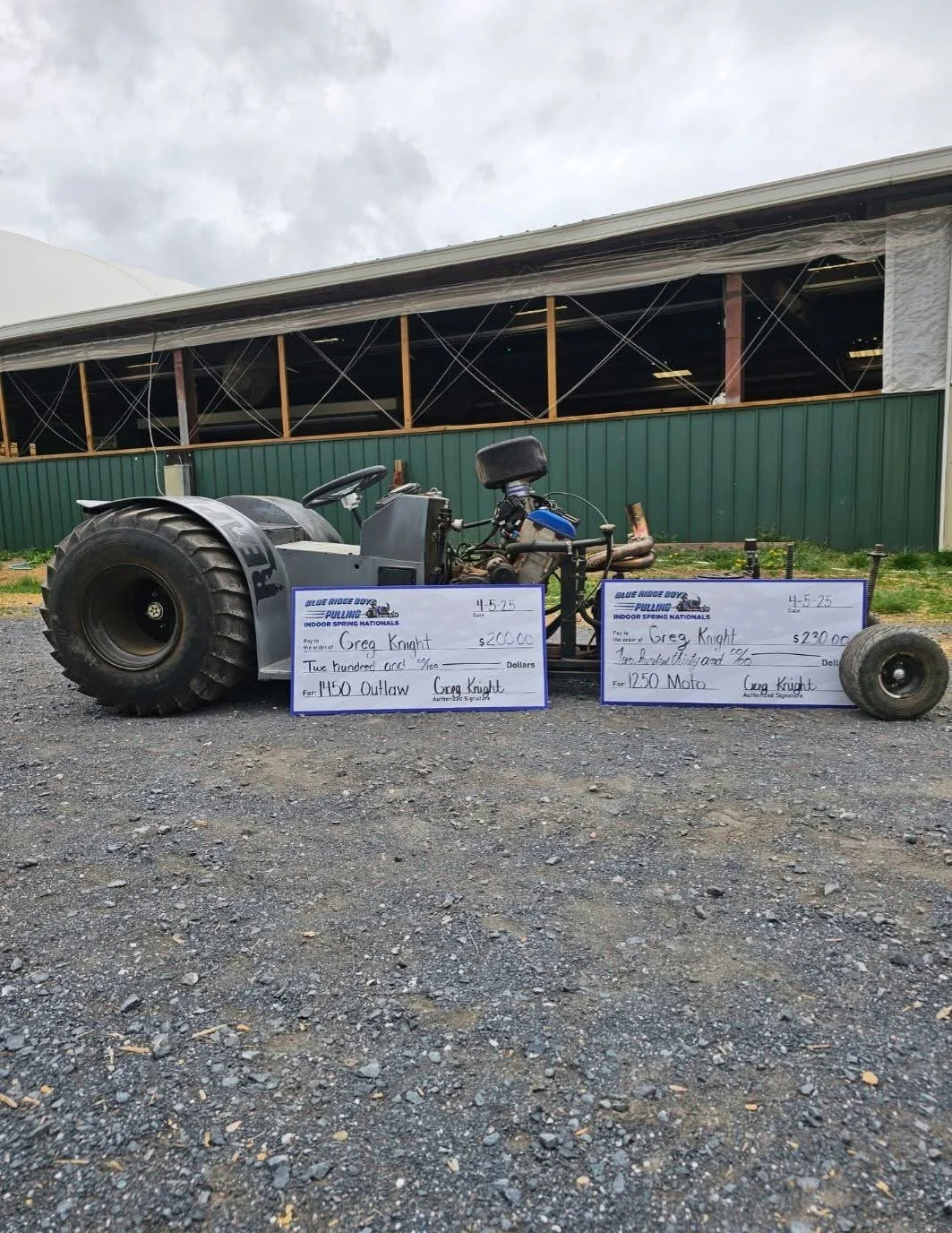 A modified racing go-kart with large rear tires and a smaller front end, parked on gravel, with large check displays for $2,000 and $2,300 in front of it, on a cloudy day outside a building under construction.