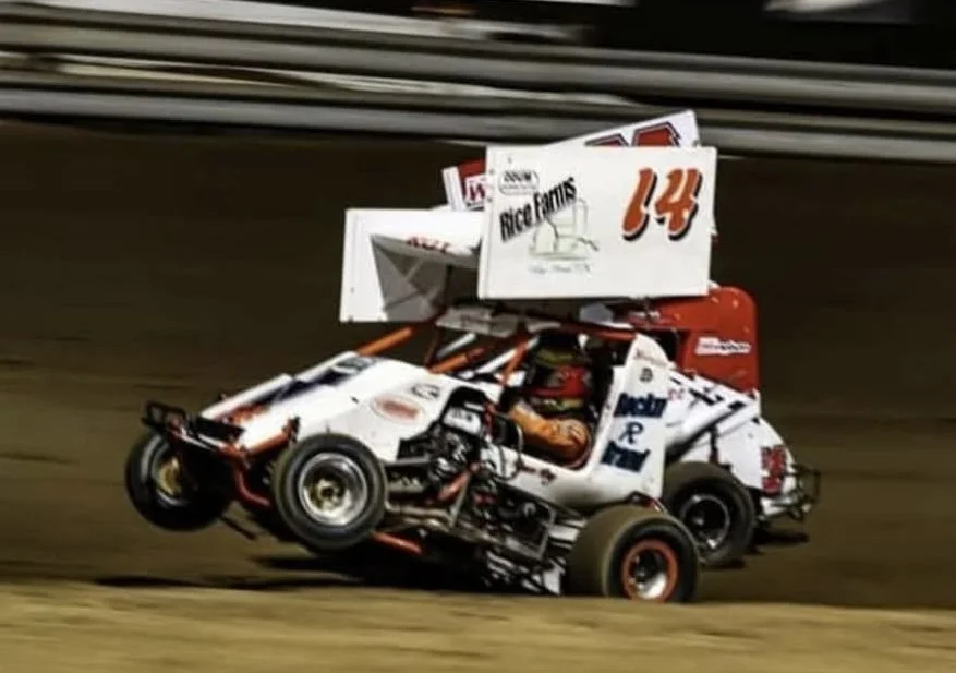 A sprint car racing on a dirt track with a large wing on top displaying the number 14.