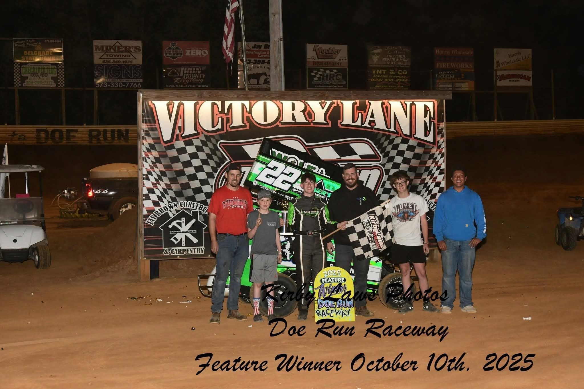 Group of six people standing in front of a racing banner at Doe Run Raceway, celebrating a victory on October 10th, 2025. The person in the center holds a checkered flag, and another holds a race car steering wheel and a winning sign.