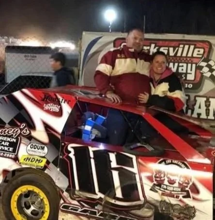 Two individuals in racing suits standing next to a red race car with the number 16, at a racing event at night.