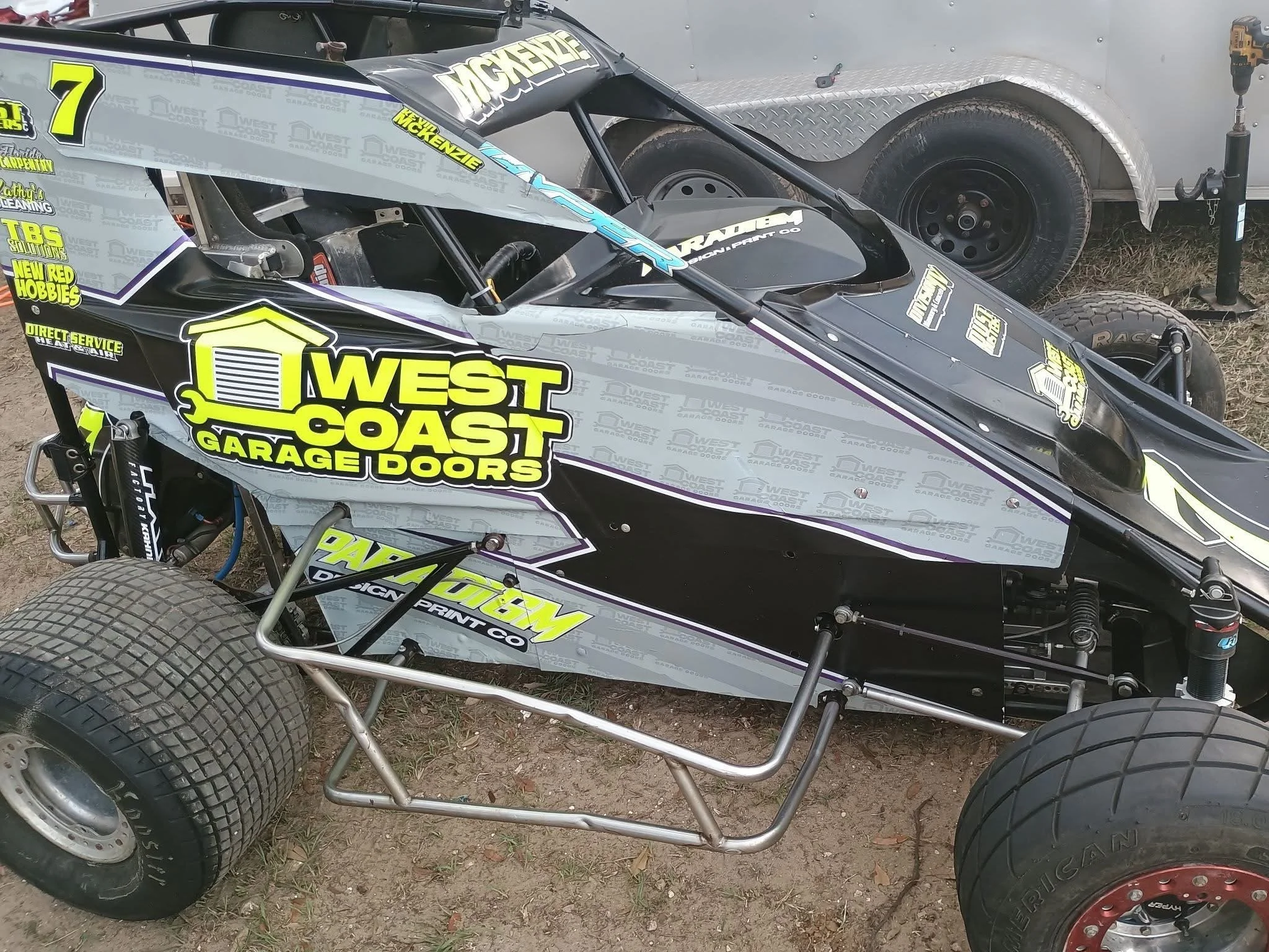 A racing car with black, grey, and yellow branding, featuring sponsor logos such as 'West Coast Garage Doors' and the number 7, parked on dirt ground near a silver trailer.