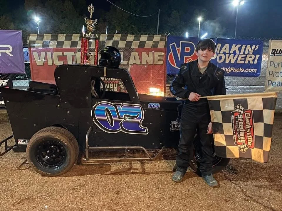 A young race car driver holding a checkered flag with the Dirt Nashville Speedway logo, standing next to a black race car number 20 with a large trophy on top, at night at a race track.