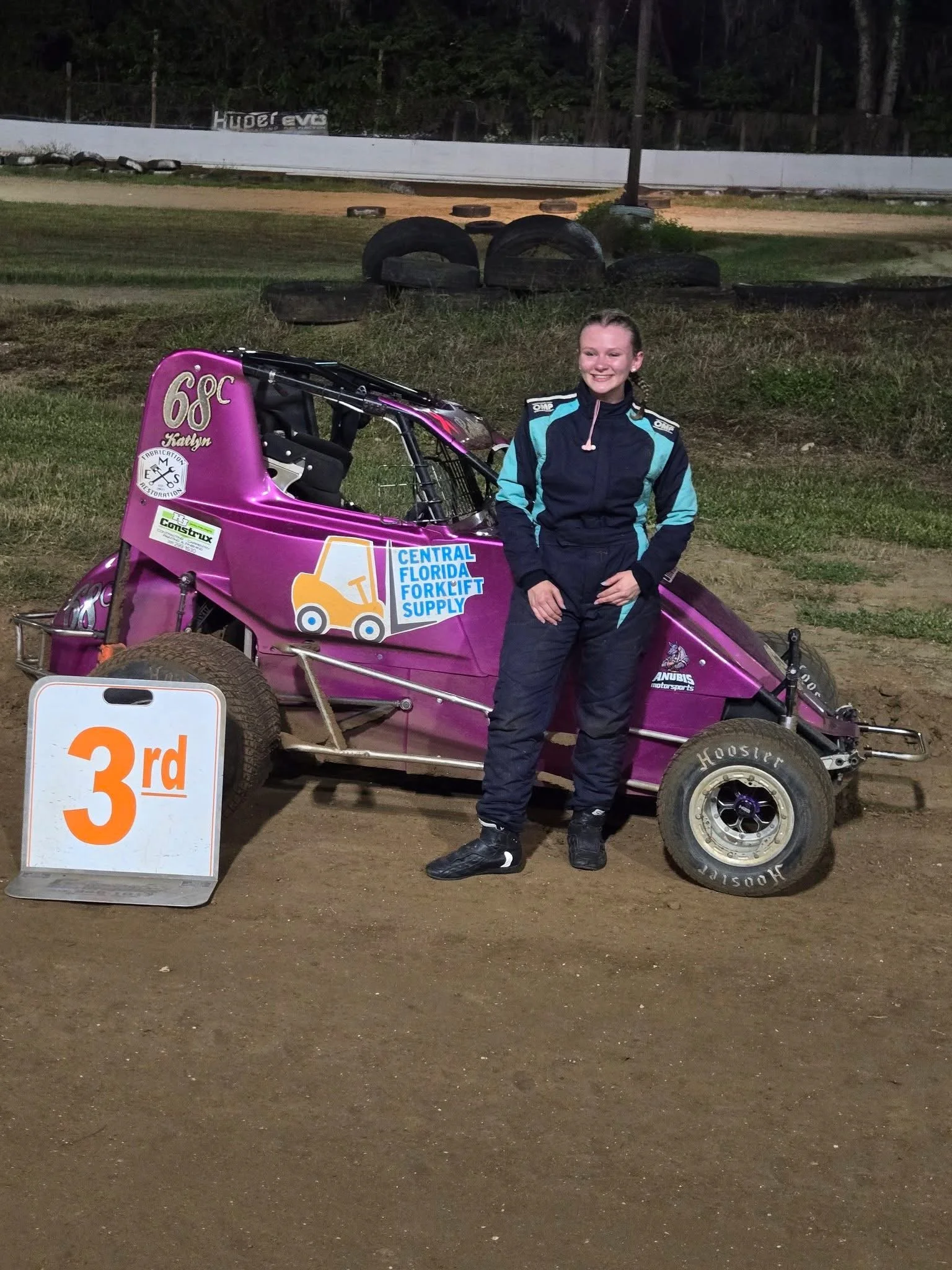 A young woman in a racing suit standing next to a purple sprint car on a dirt race track, with a sign indicating third place finish.