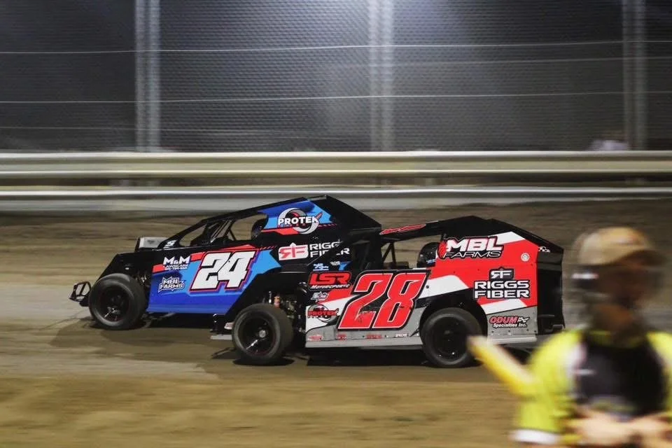 Two sprint cars racing on a dirt track, one black and blue with the number 24, the other black and red with the number 28, with a person in a yellow vest in the foreground.