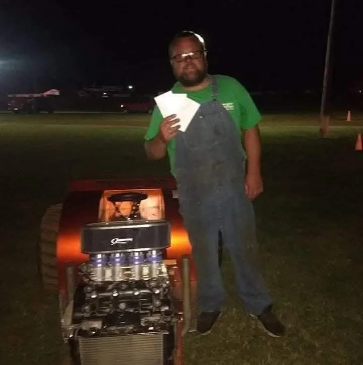 Man standing outdoors at night, holding papers, next to small custom go-kart with engine and small tracks.