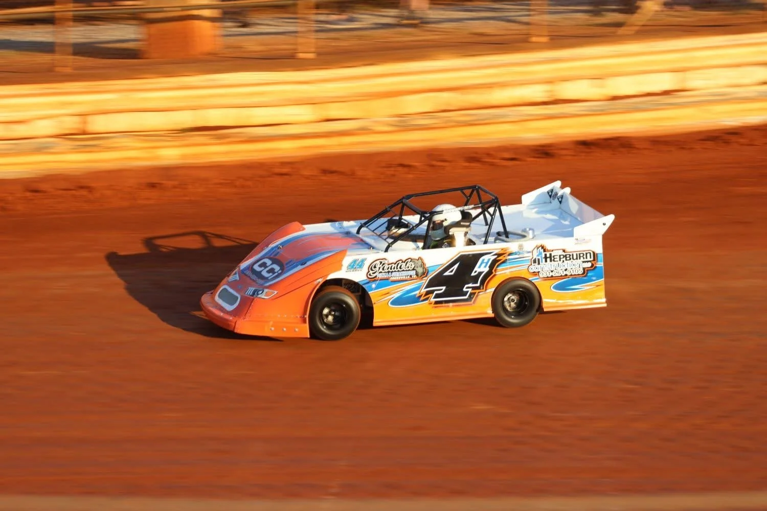 An orange, blue, and white race car with the number 4 on its side speeding on a dirt track during a race.