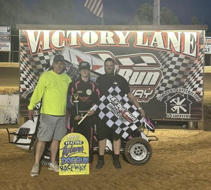 Three men standing together at a raceway, one holding a checkered flag, celebrating a victory at the 2025 Doe Run Raceway, with a sprint car behind them and a large banner reading 'Victory Lane' in the background.