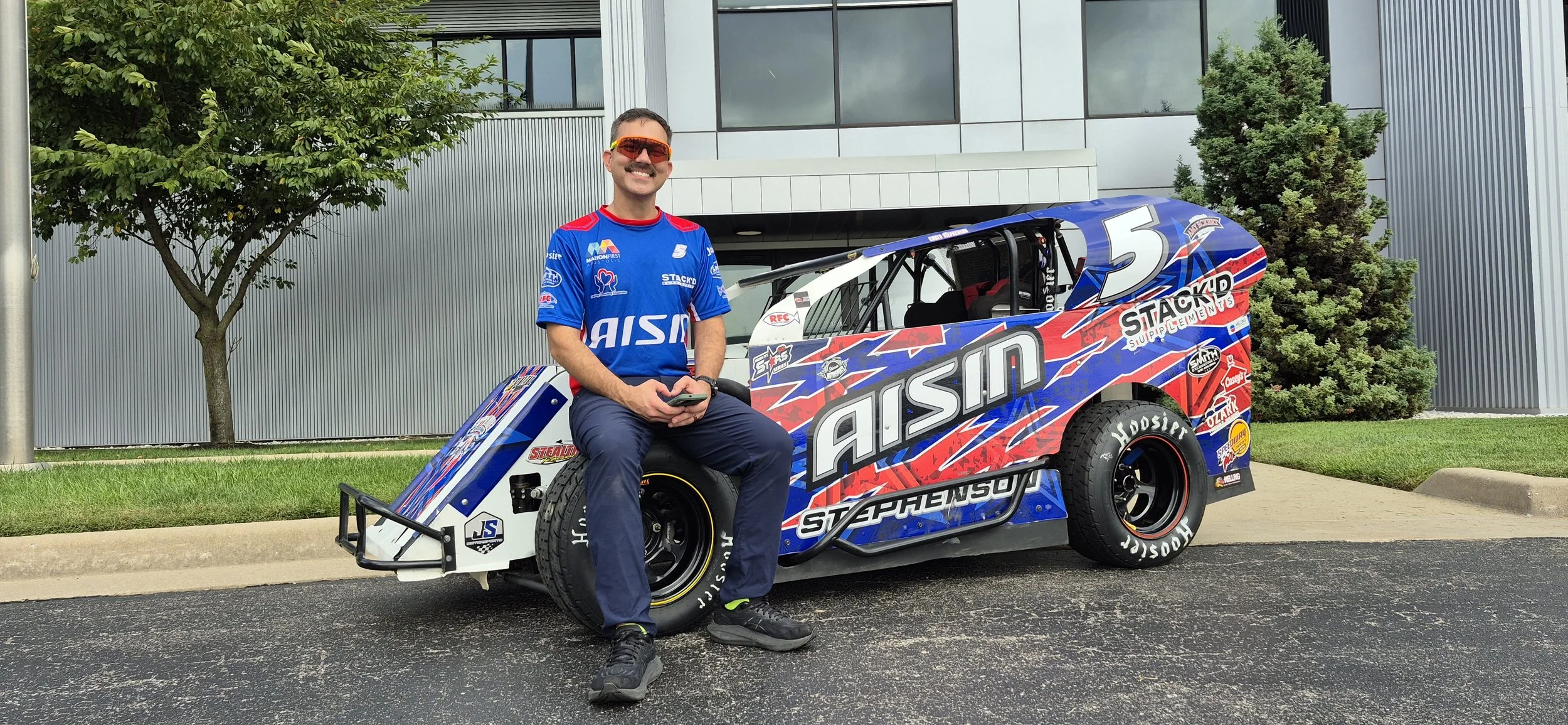 A man in a blue racing shirt and jeans sitting on a race car, smiling, beside a modern building with trees in the background.