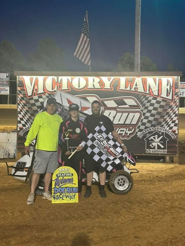 Three individuals stand on a dirt raceway in front of a large Victory Lane banner after a race. The person in the middle is holding a trophy and a checkered flag. A small race car is visible behind them, and the scene is illuminated with floodlights.