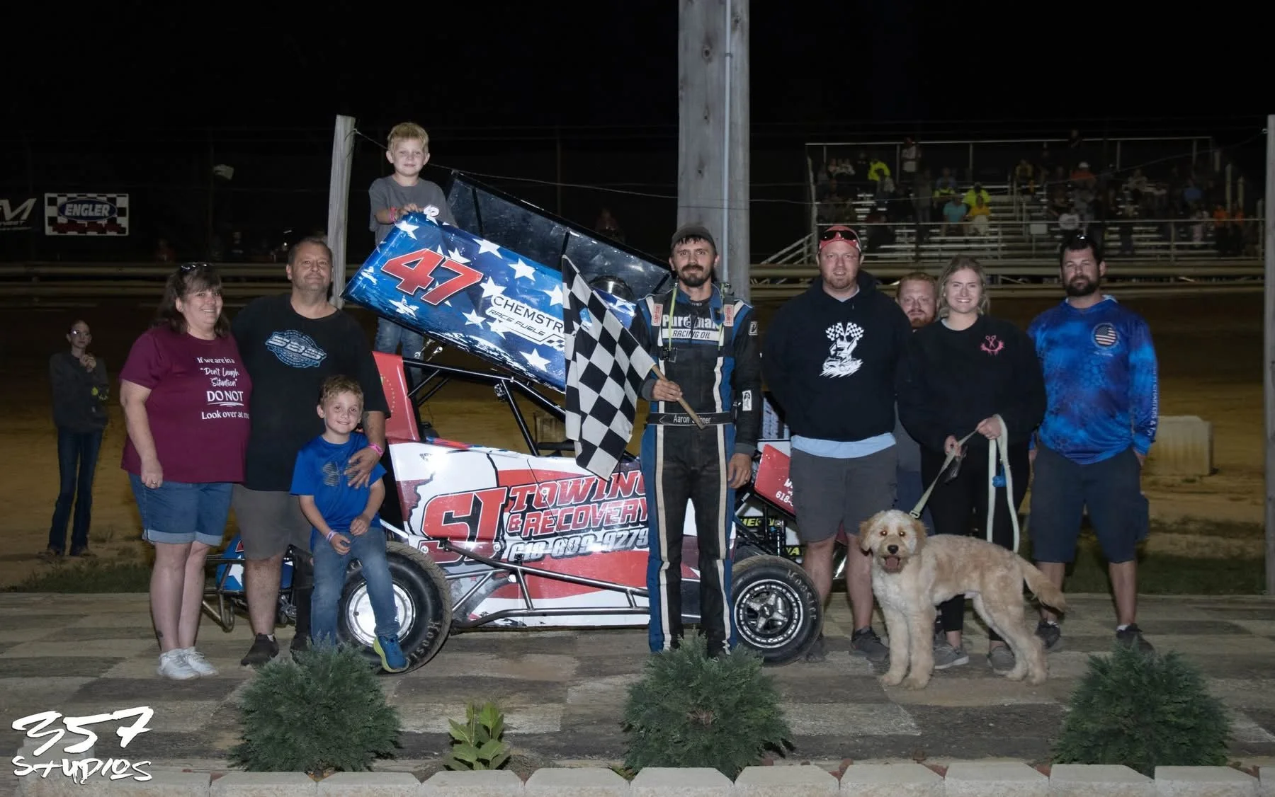 Group of people, including children and adults, posing with a racing car and a dog at a nighttime racing event, with spectators in the background.