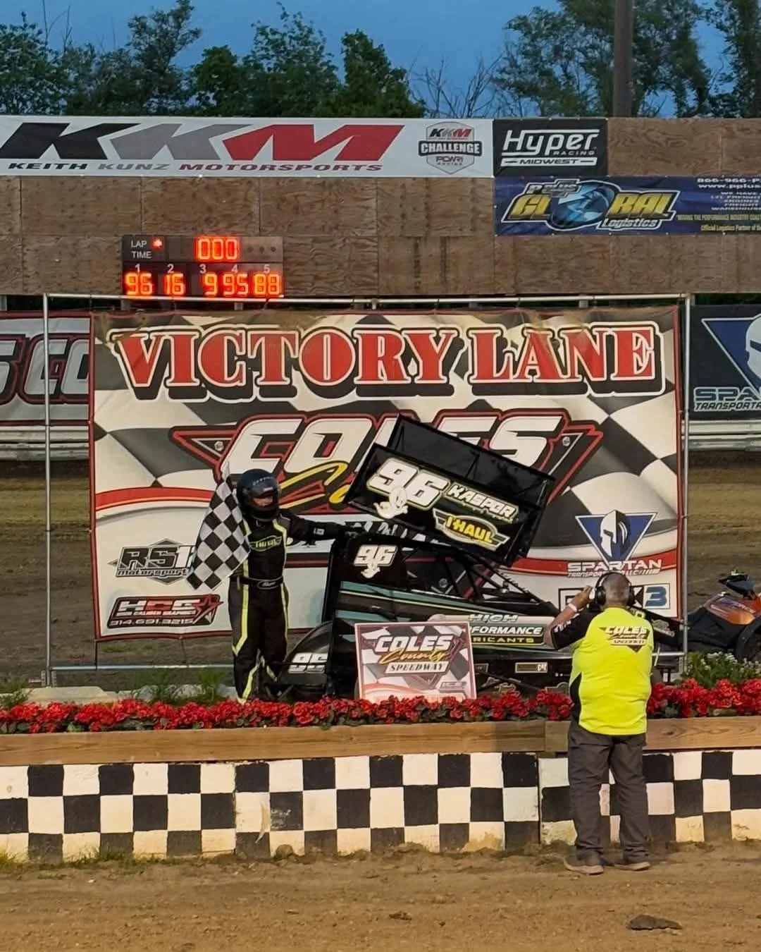 Race car driver holding checkered flag next to his sprint car on the winner's podium at a dirt race track, with a person taking a photo. The background shows banners and a digital scoreboard with the race results.