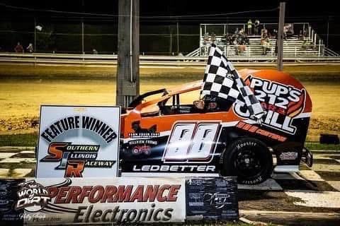 Orange race car with the number 66, checkered flag, and sponsorship logos, situated on a dirt track with banners and bleachers in the background.