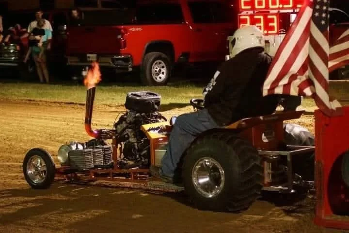 A person wearing a helmet and black jacket sitting on a small, custom-built tractor pulling a wagon with an American flag at a dirt race track during nighttime. In the background, there are people and a red truck, with bright digital scoreboard displ