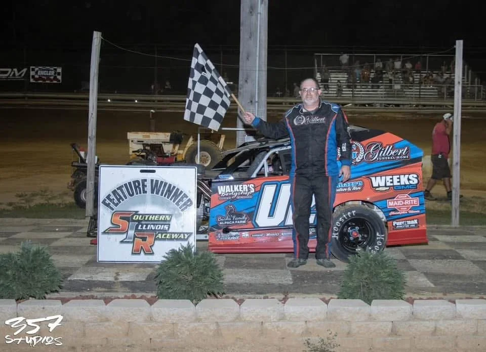 A man in racing overalls holding a checkered flag, standing next to a race car with the number 07, at a dirt race track during night time. There is a sign in front that says 'Feature Winner Southern Illinois Raceway.'