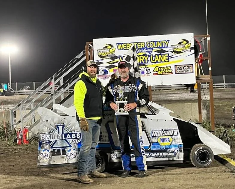 Two men standing in front of a race car at Webster County Speedway, one holding a trophy, with a checkered flag behind them and a sign for the race lane in the background.