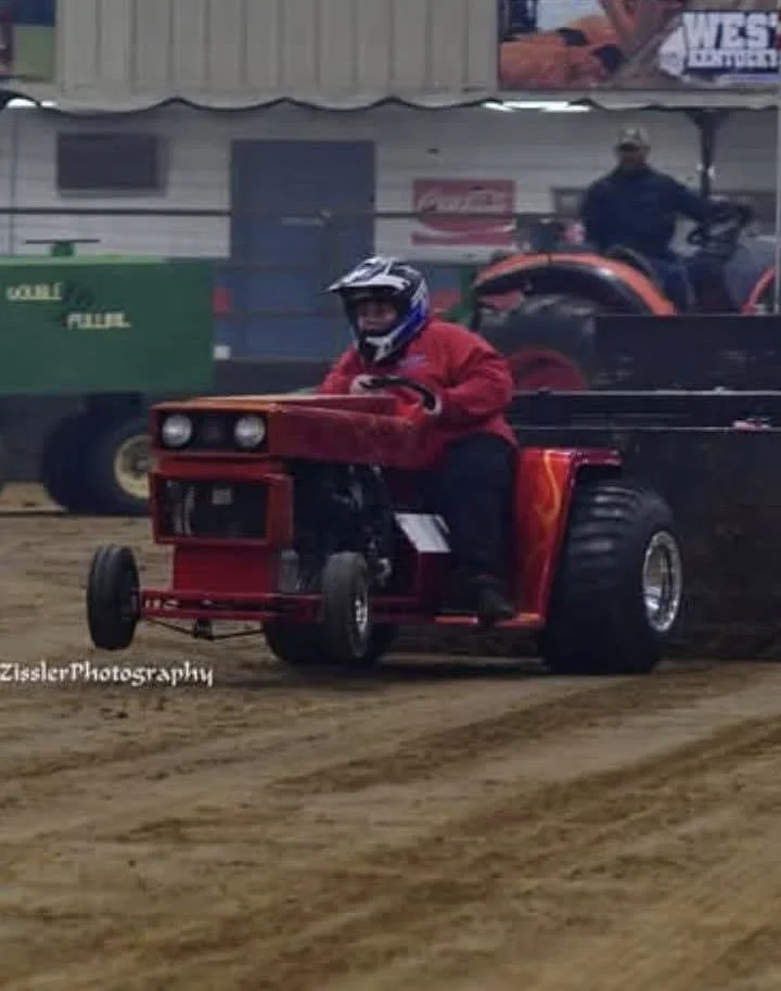 A person wearing a red jacket and helmet driving a small, modified, red tractor on a dirt track at a racing event, with another person on an ATV in the background.