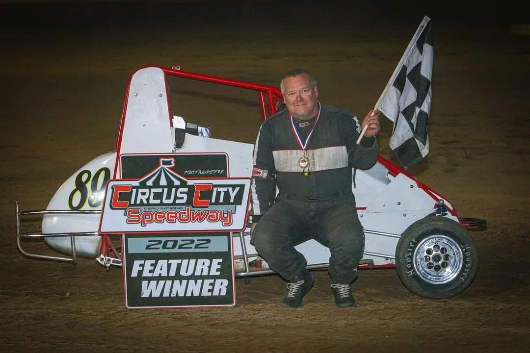 A man kneeling next to a small race car holding a checkered flag at a speedway track. There is a sign that says 'CircuCity Speedway 2022 Feature Winner' in front of him.